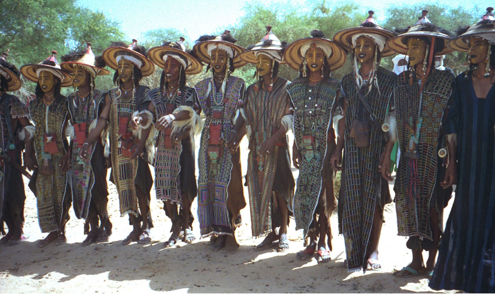 Fila de hombres con sombreros cónicos de paja, rostros pintados de amarillo y túnicas bordadas, bailando hombro con hombro sobre la arena.