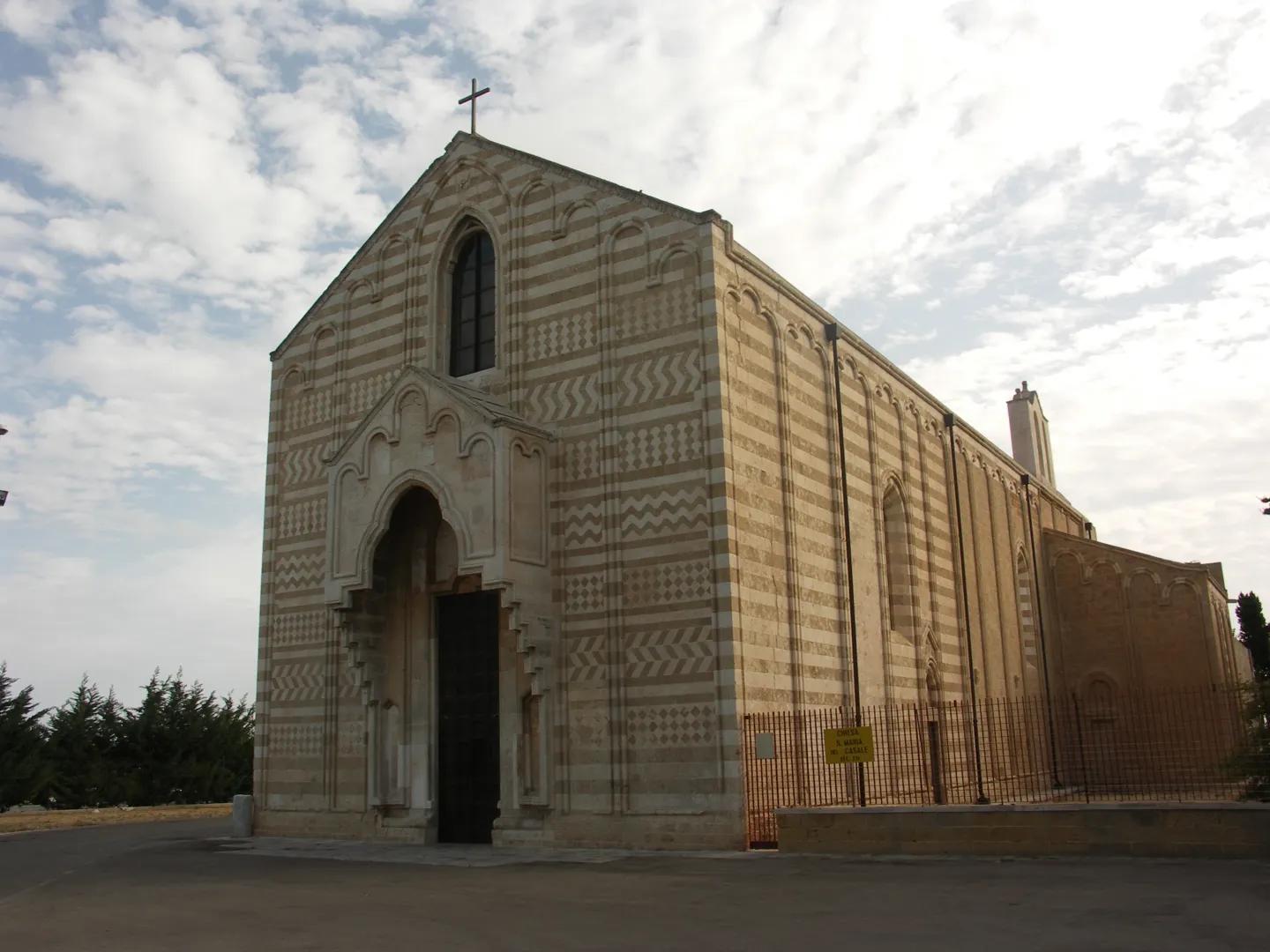 Fachada de la catedral de Brindisi con piedras alternas beige y blancas, arco ojival y puerta de madera, bajo un cielo nublado con árboles al fondo