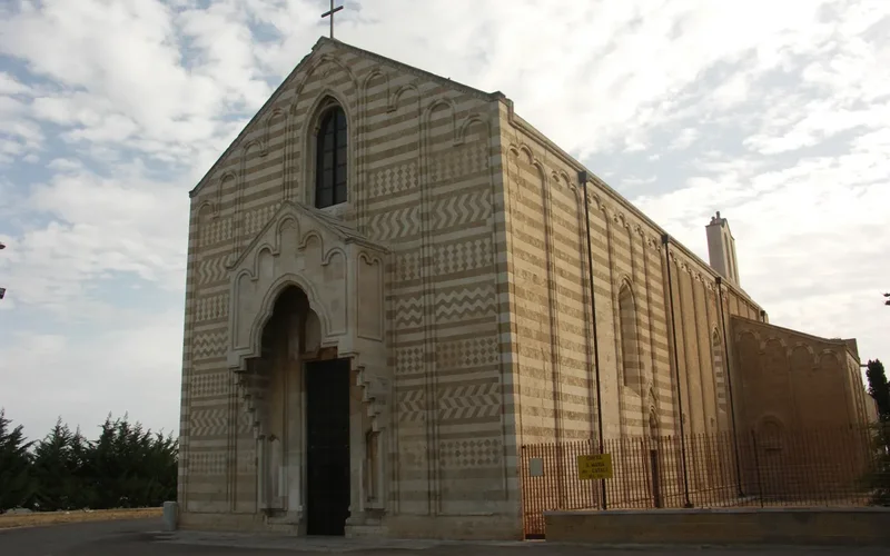 Fachada de la catedral de Brindisi con piedras alternas beige y blancas, arco ojival y puerta de madera, bajo un cielo nublado con árboles al fondo