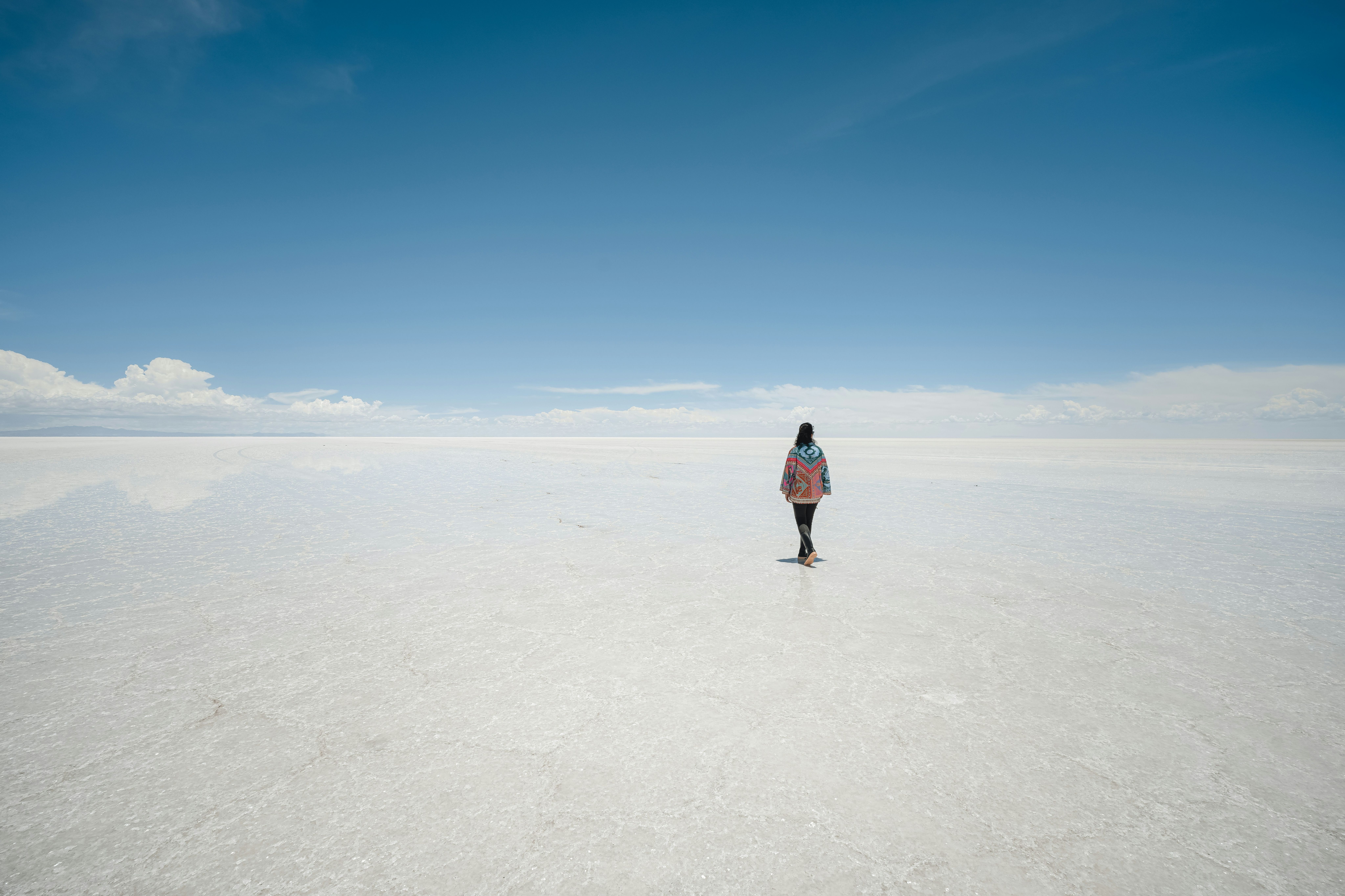 Una persona de espaldas caminando sobre una vasta superficie blanca que refleja el cielo azul.