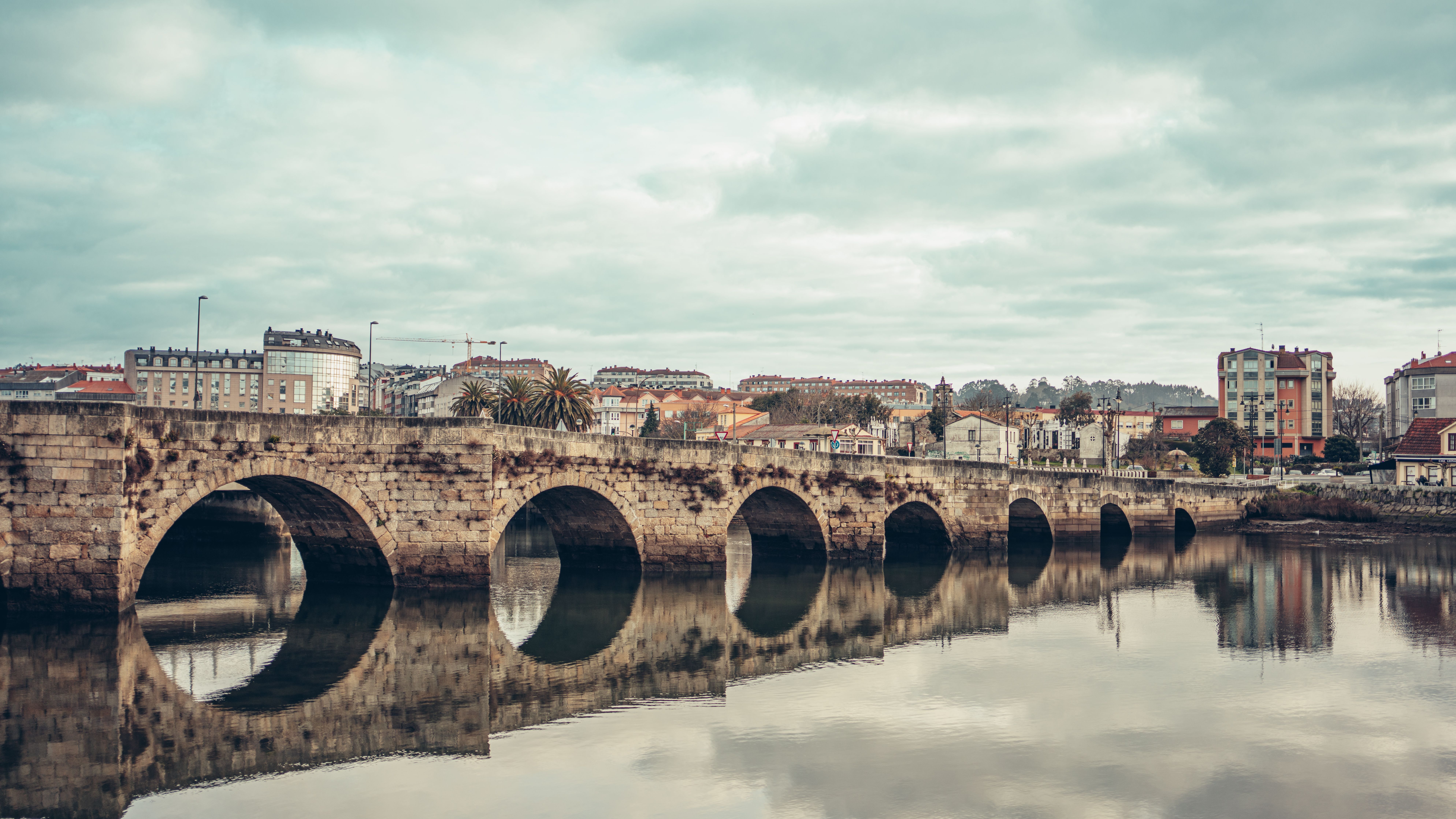 Puente de piedra con arcos sobre un río, reflejándose en el agua, con edificios al fondo bajo un cielo nublado.