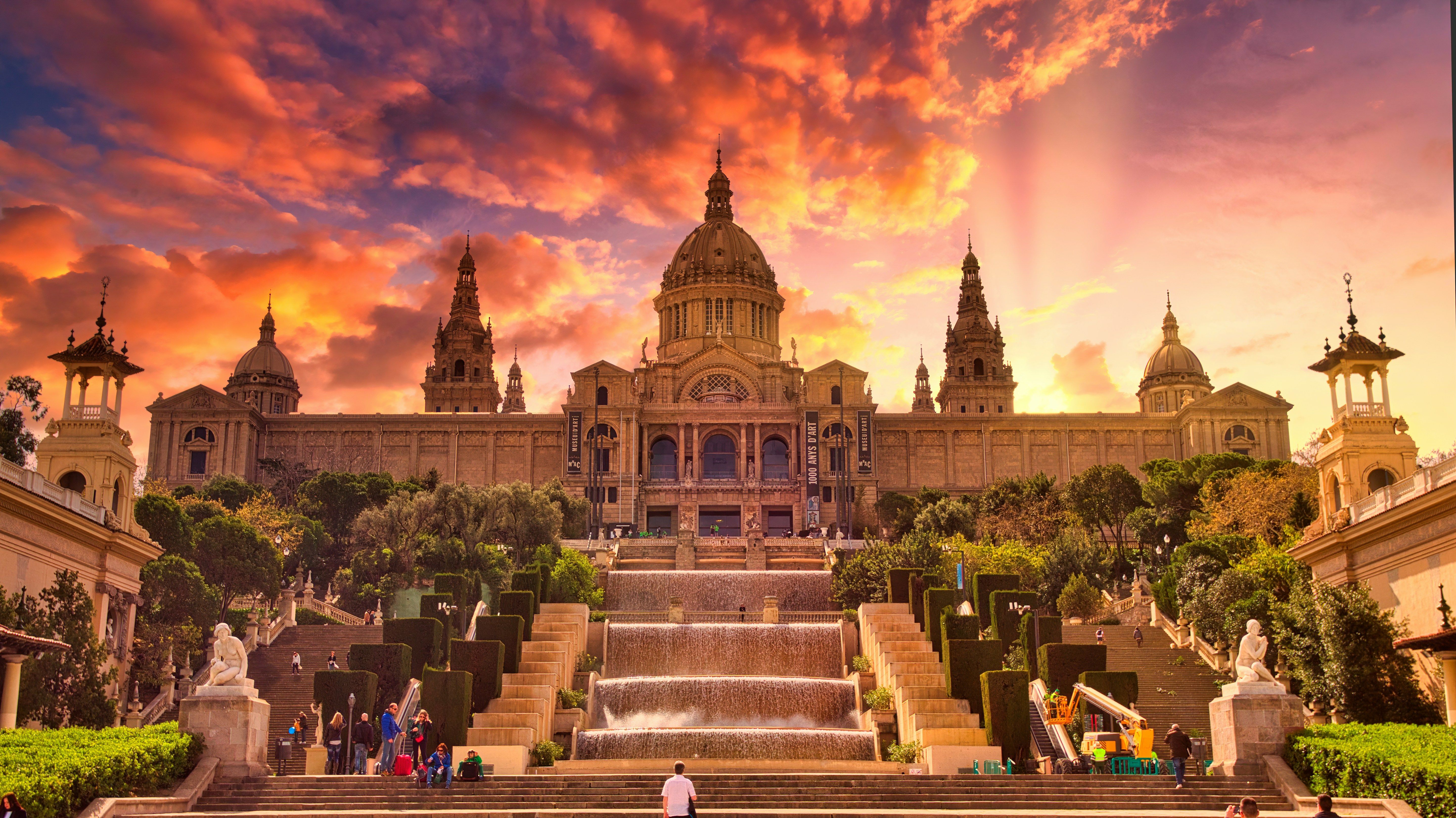 Vista del Palacio Nacional de Barcelona al atardecer, con escalinatas, cascada y jardines, bajo un cielo rosado y naranja