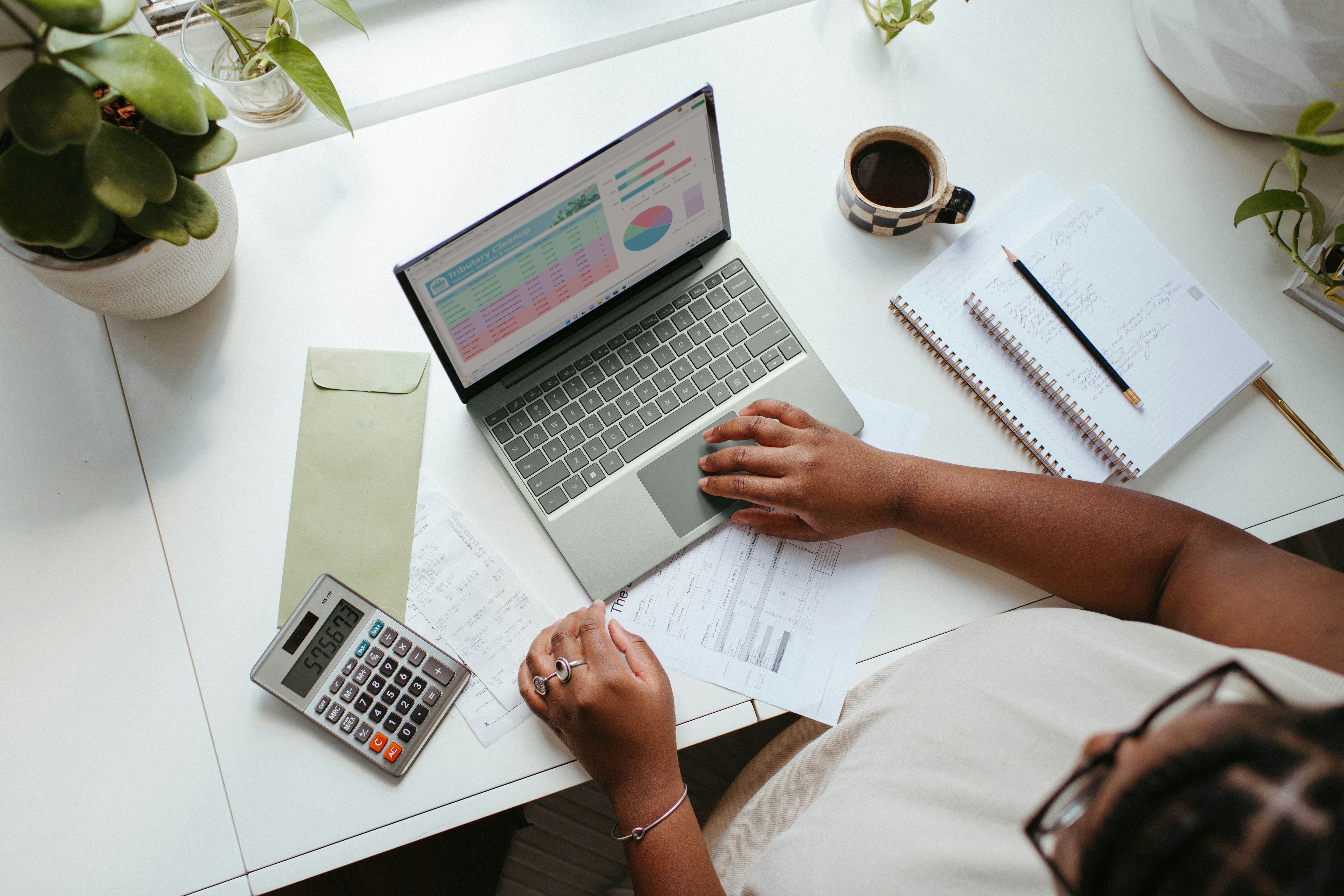 Persona trabajando en un portátil con calculadora, documentos y café sobre un escritorio blanco.