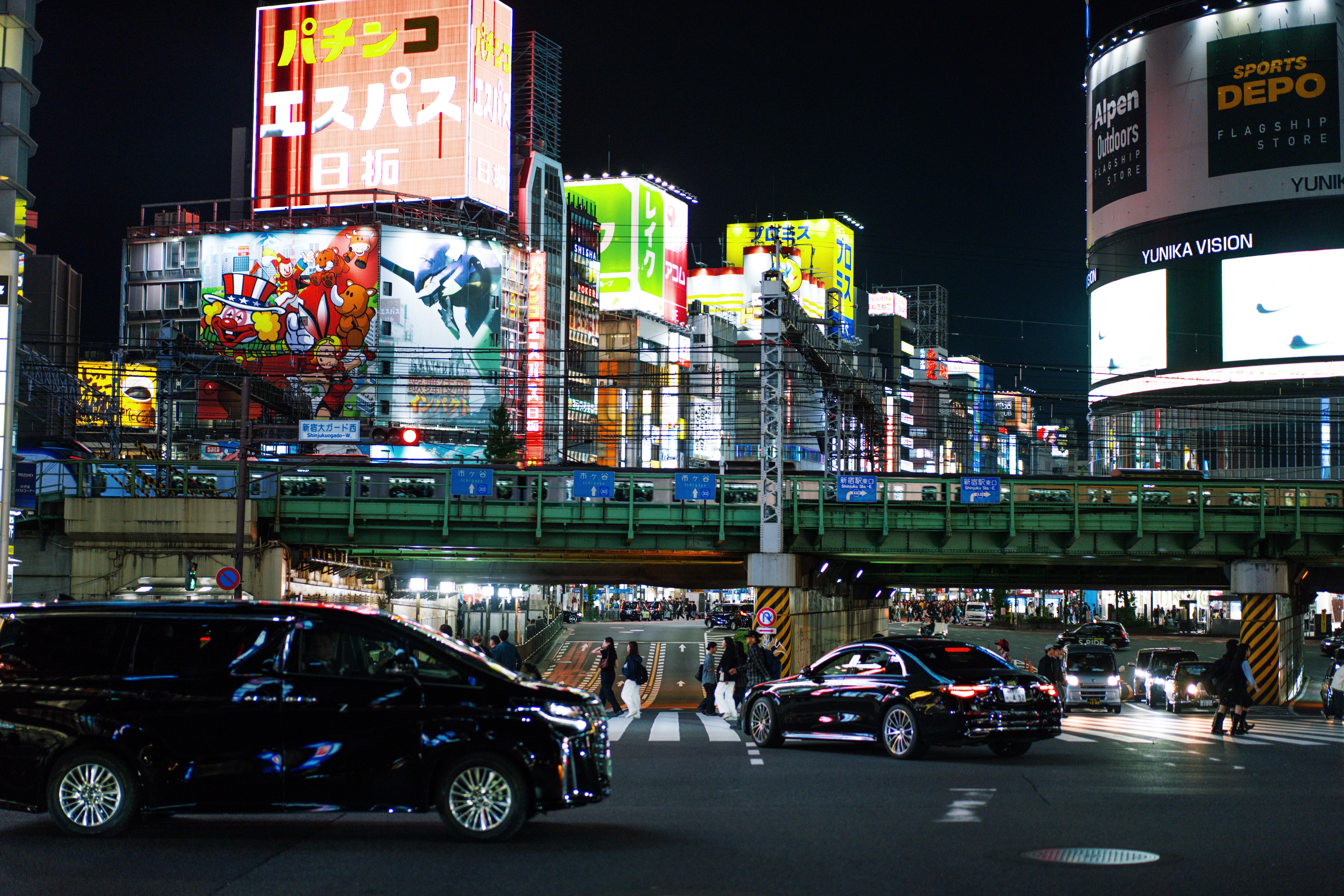 Vista nocturna de una calle con coches y personas, edificios iluminados con carteles publicitarios en japonés.