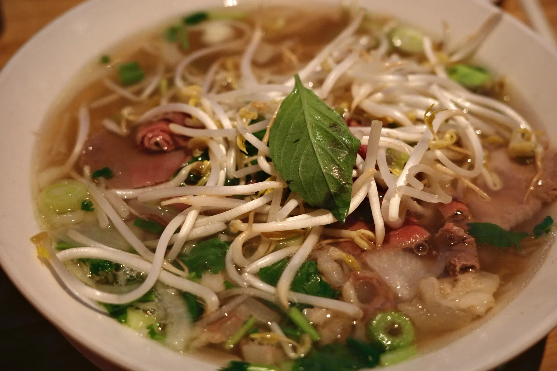 Un tazón blanco lleno de sopa pho con fideos, brotes de soja, carne y una hoja verde.
