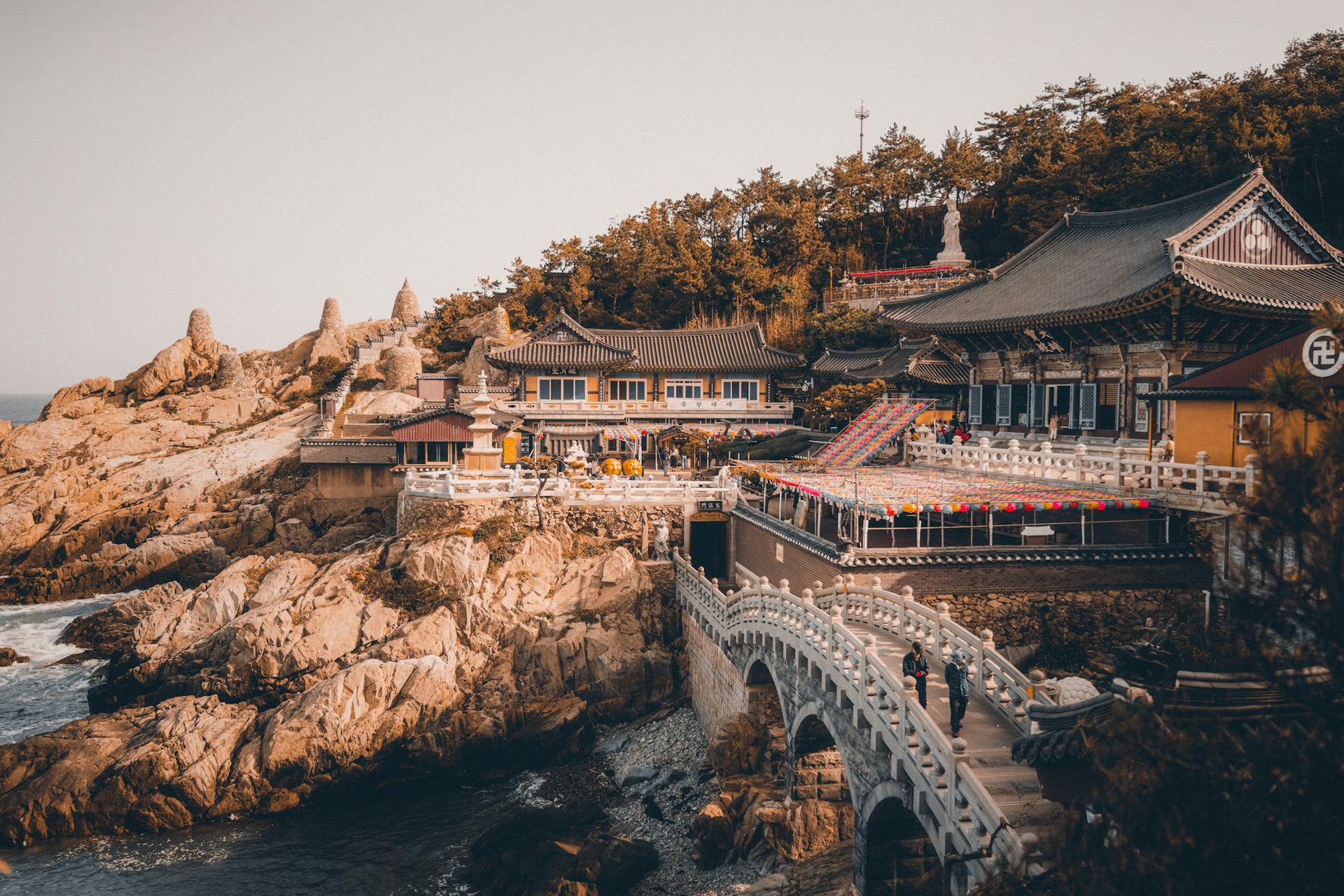 Vista panorámica de templo budista en acantilado rocoso de Busan, con puente blanco arqueado, faroles rojos y edificios tradicionales junto al mar