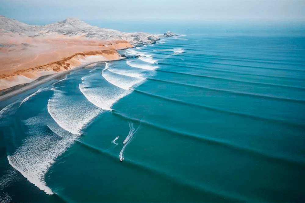 Vista aérea de la costa de Puerto Chicama con olas largas rompiendo en paralelo junto a dunas desérticas y un surfista en el agua