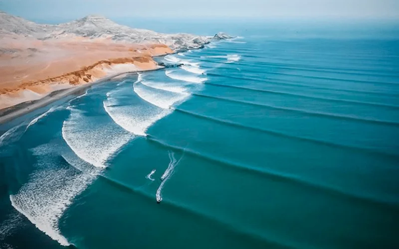 Vista aérea de la costa de Puerto Chicama con olas largas rompiendo en paralelo junto a dunas desérticas y un surfista en el agua