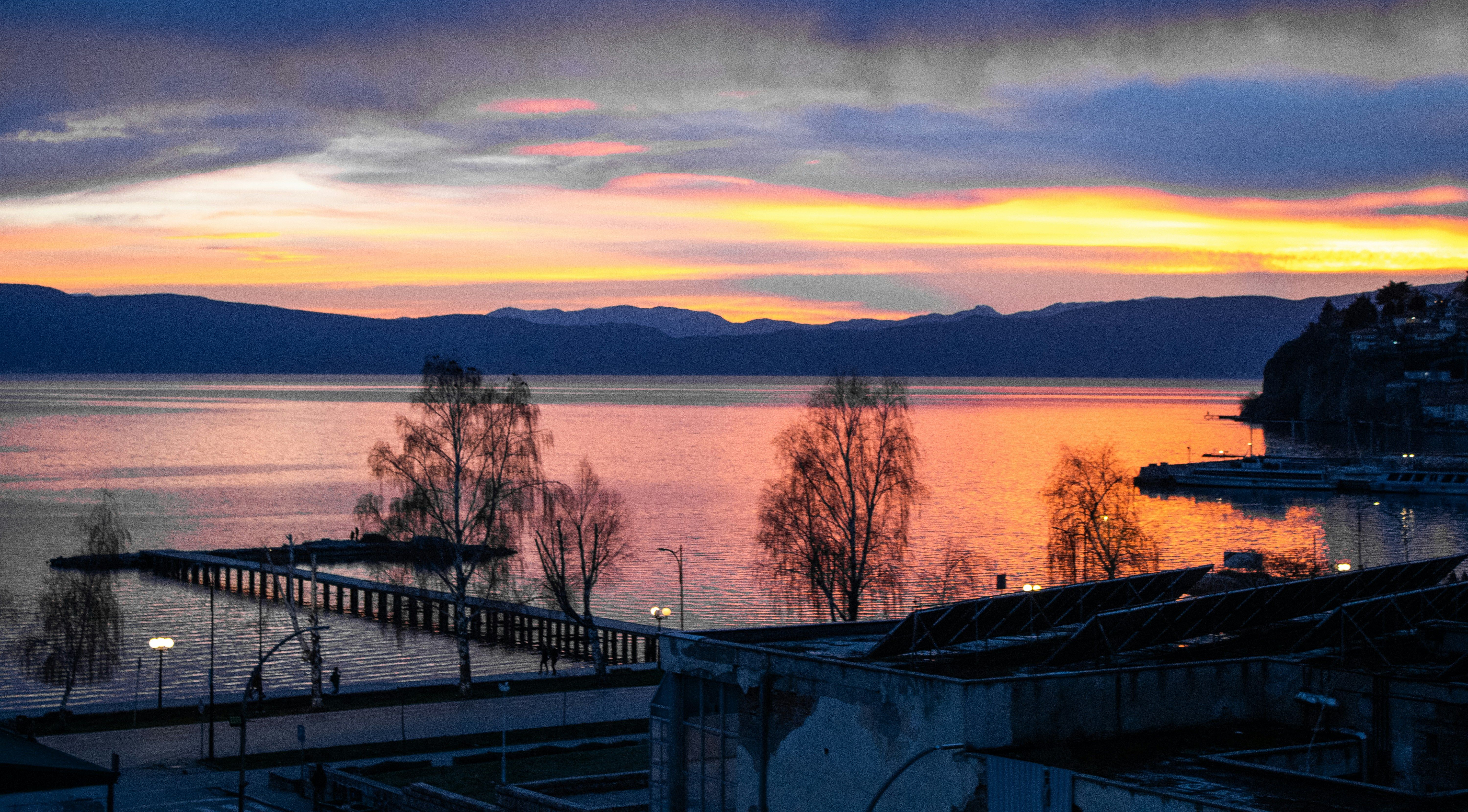 Vista panorámica del lago Ohrid al amanecer con un muelle, árboles y montañas en el horizonte.
