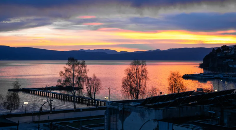 Vista panorámica del lago Ohrid al amanecer con un muelle, árboles y montañas en el horizonte.
