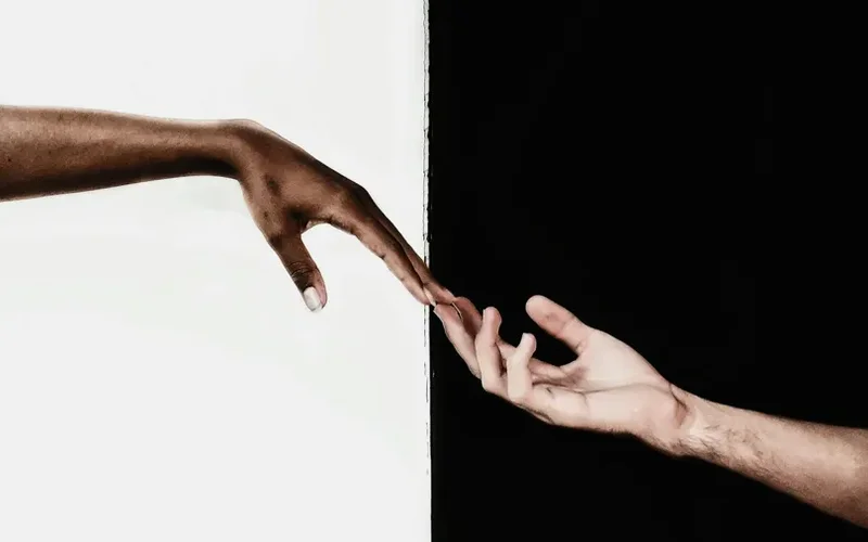 Dos manos de tonos de piel distintos se acercan casi tocándose, divididas por una línea vertical en un fondo mitad blanco y mitad negro