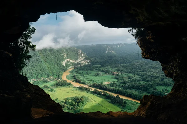 Vista de un valle verde con un río marrón serpenteante, enmarcado por la entrada oscura de una cueva.
