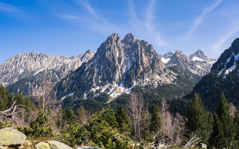 Montañas rocosas de los Pirineos con nieve en las cumbres, picos afilados bajo cielo azul y bosque de coníferas en primer plano