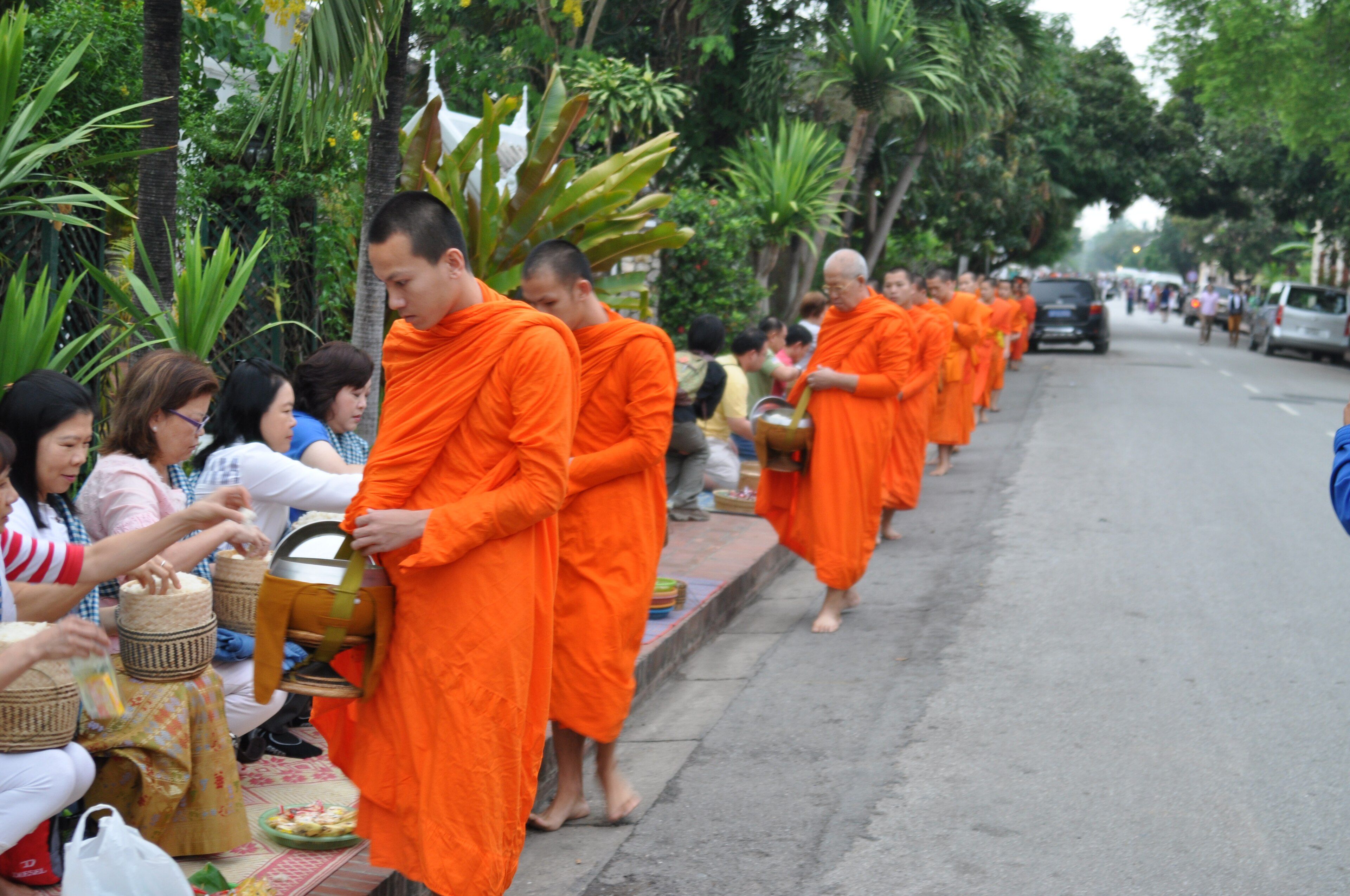 Fila de monjes con túnicas naranjas y cuencos que caminan descalzos por una acera mientras mujeres sentadas les ofrecen comida de cestas.
