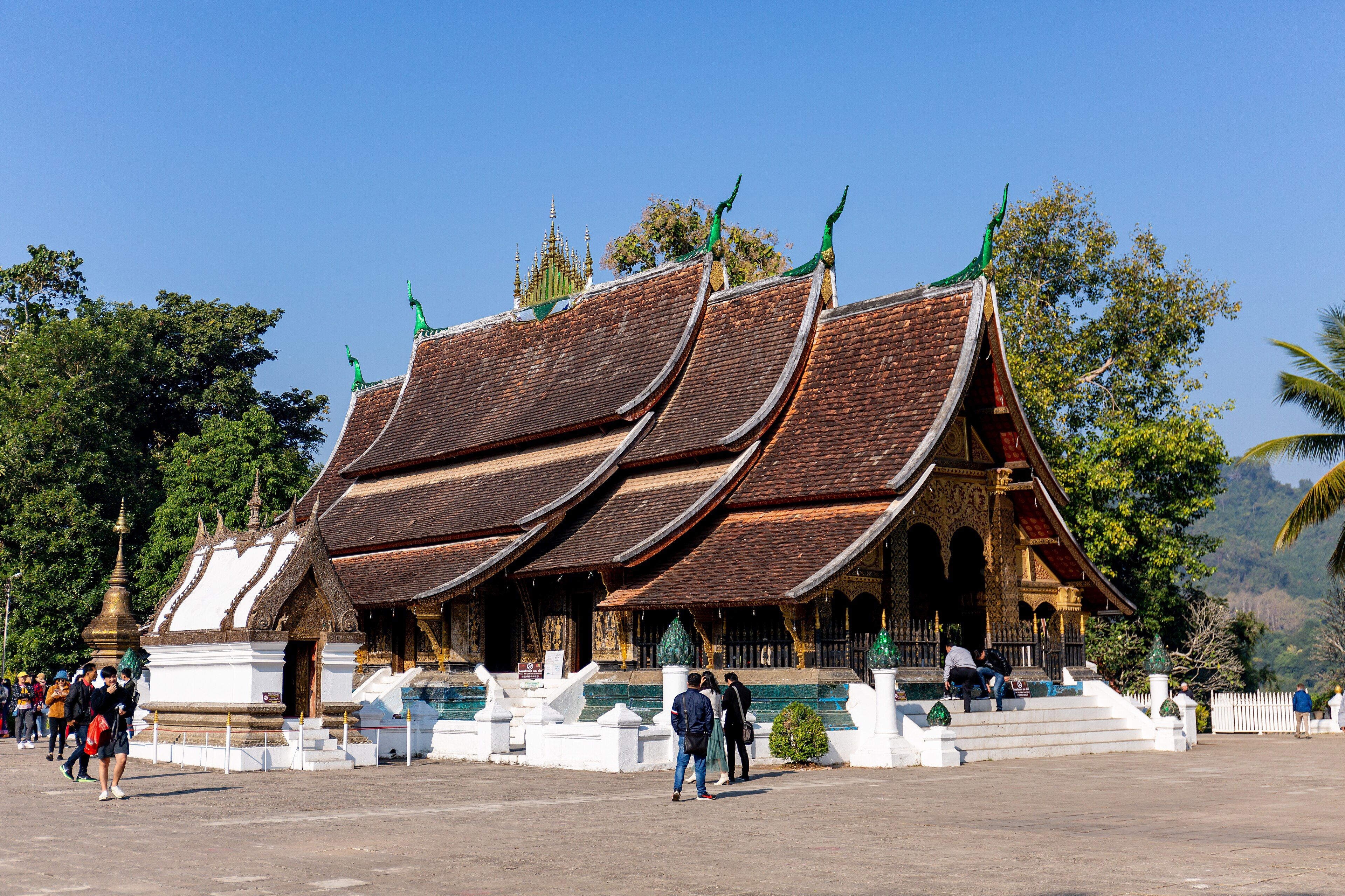 Templo budista de tejados oscuros superpuestos que bajan casi hasta el suelo, con remates verdes y dorados, en una explanada bajo un cielo azul.