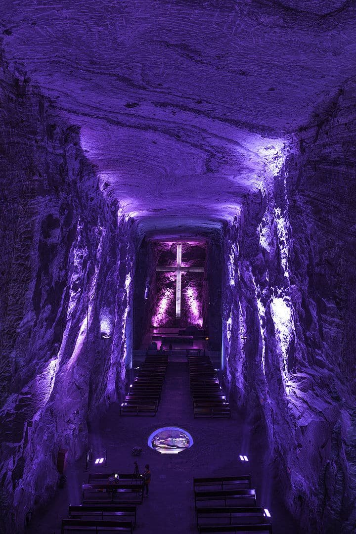 Interior de la Catedral de Sal de Zipaquirá iluminado en tonos púrpura, con cruz central resplandeciente y bancos alineados en el suelo rocoso