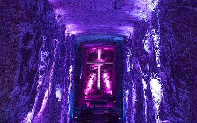 Interior de la Catedral de Sal de Zipaquirá iluminado en tonos púrpura, con cruz central resplandeciente y bancos alineados en el suelo rocoso