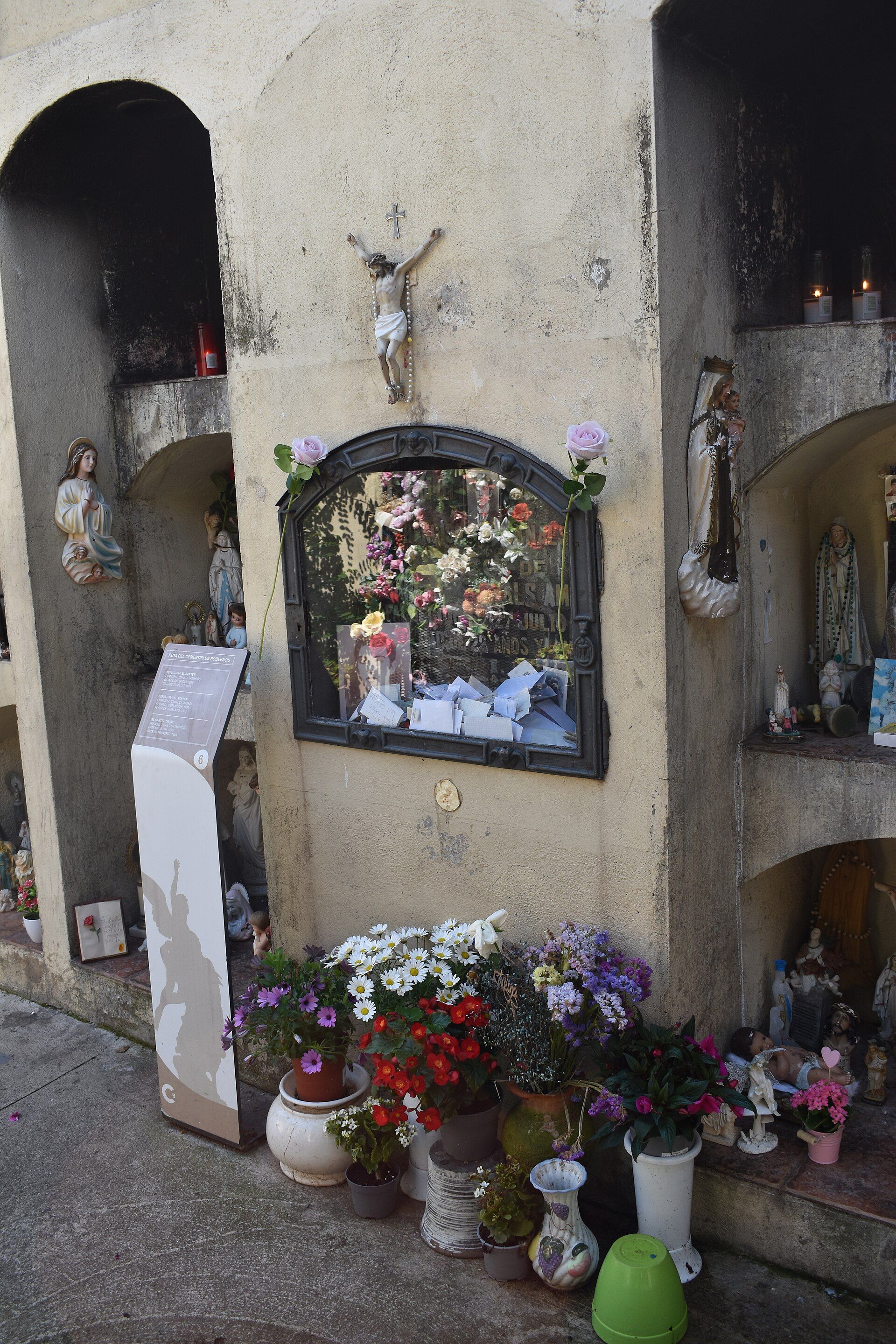 Nichos de cementerio con un crucifijo, flores, estatuas religiosas y ofrendas en un muro de piedra.