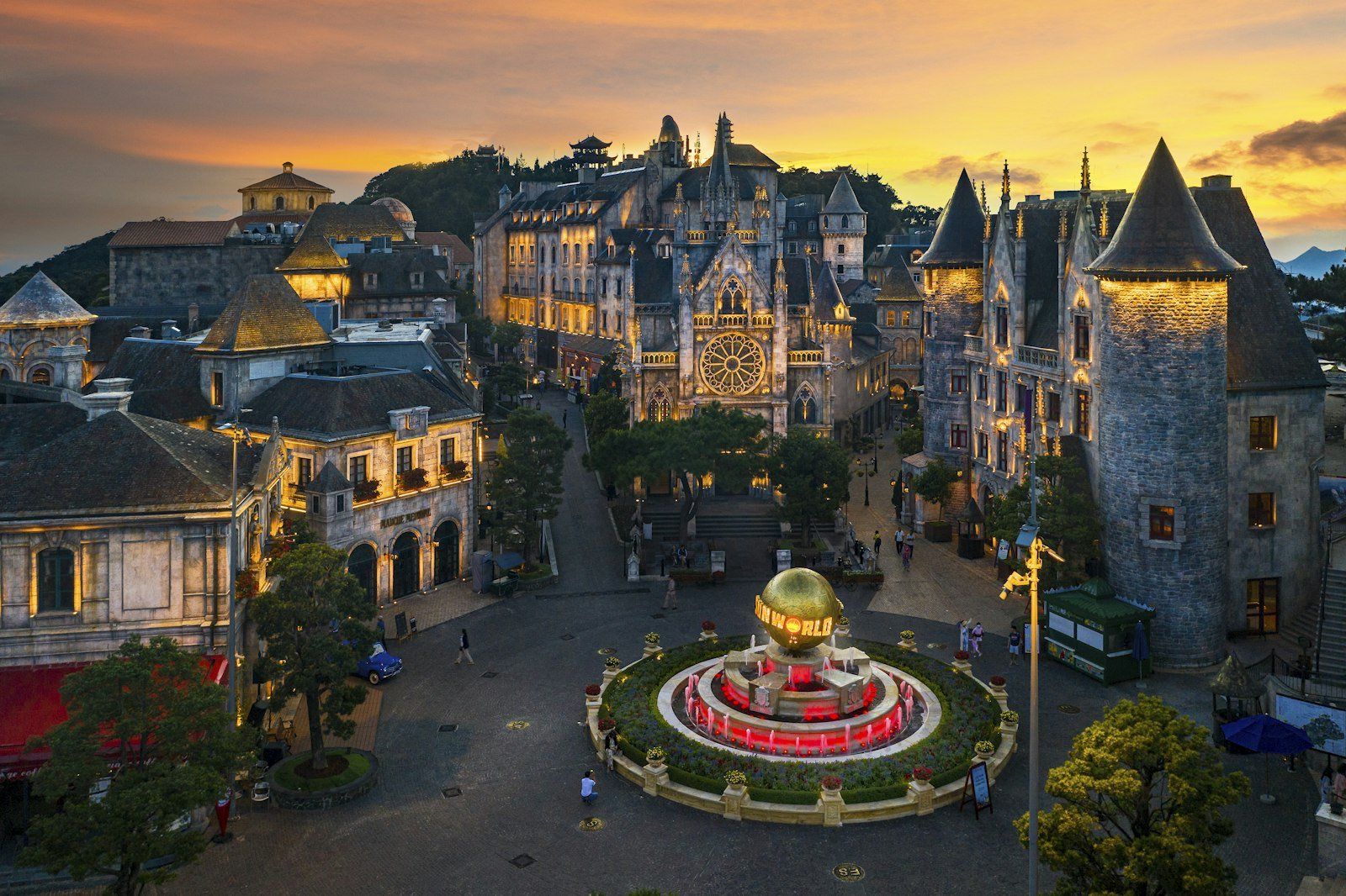 Vista al atardecer de una plaza con edificios de estilo francés, iglesia con reloj central y fuente de globo dorado rodeada de gente
