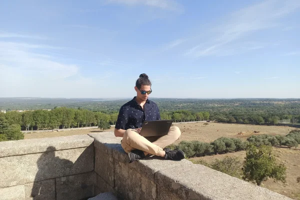 Hombre con moño y gafas de sol sentado en un muro de piedra usando un portátil, con un paisaje rural de fondo.