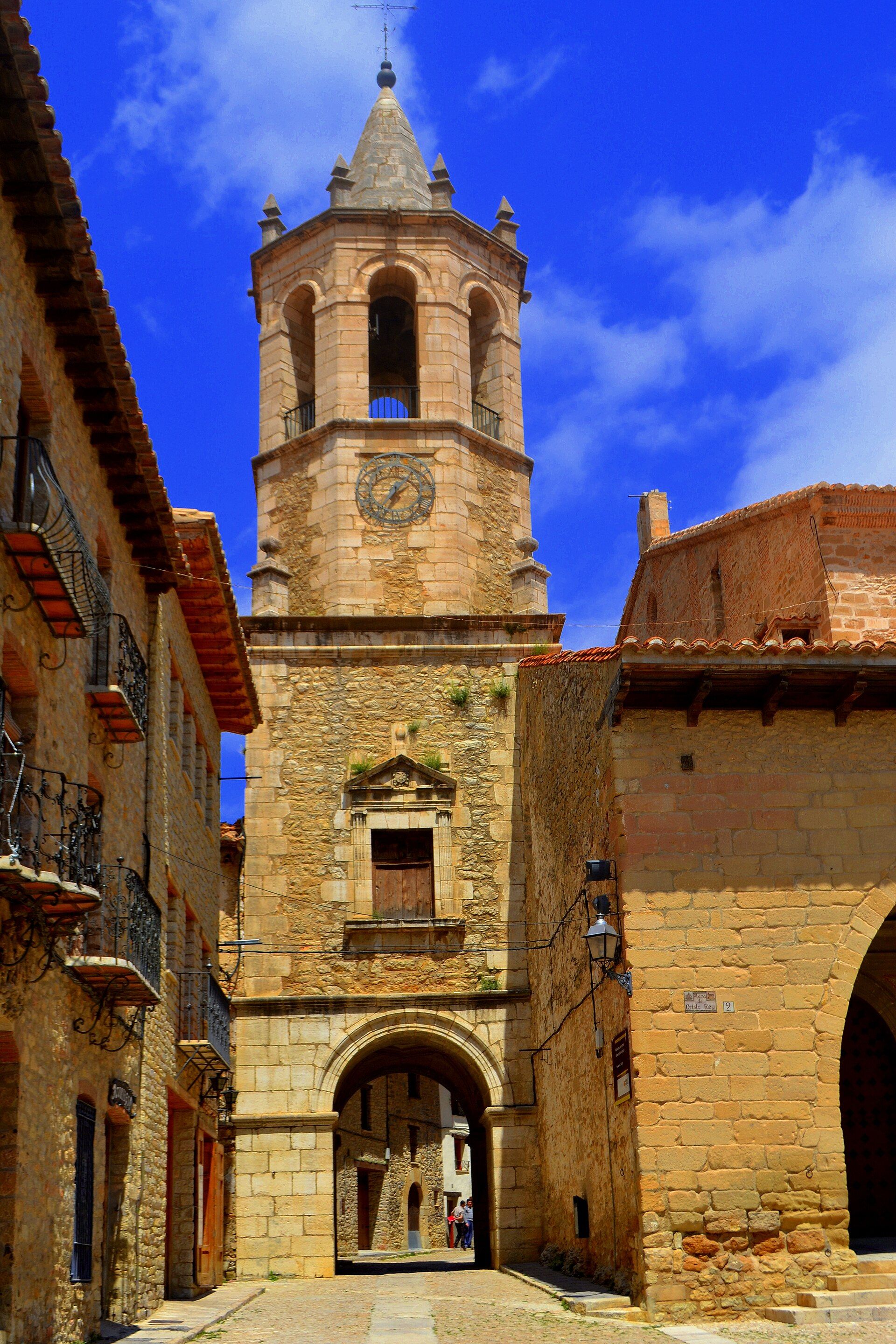 Callejón de piedra con edificios antiguos y una torre de iglesia con reloj bajo cielo azul.