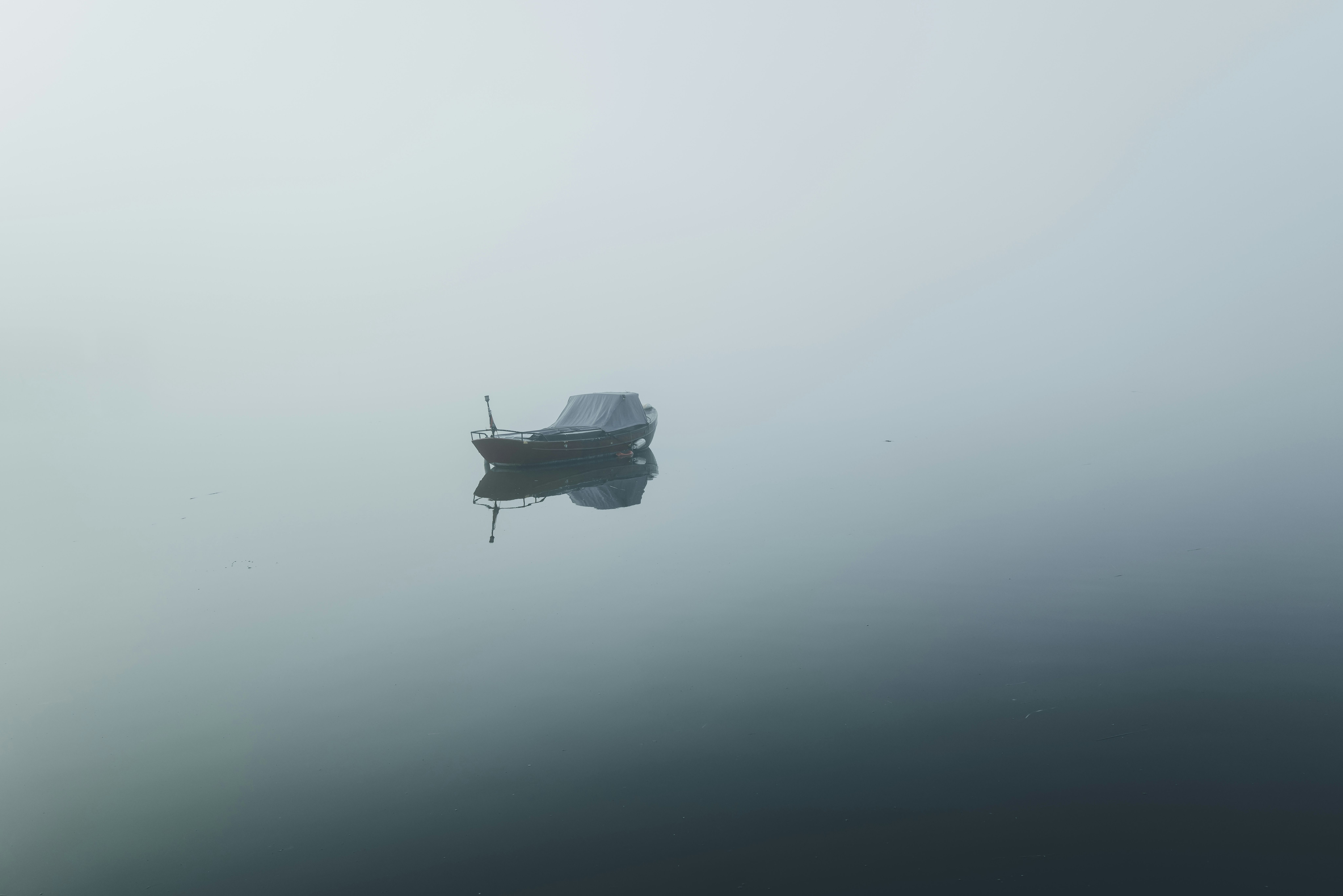 Un pequeño bote oscuro flotando en aguas tranquilas y brumosas, con su reflejo visible.