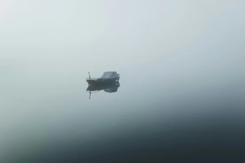 Un pequeño bote oscuro flotando en aguas tranquilas y brumosas, con su reflejo visible.