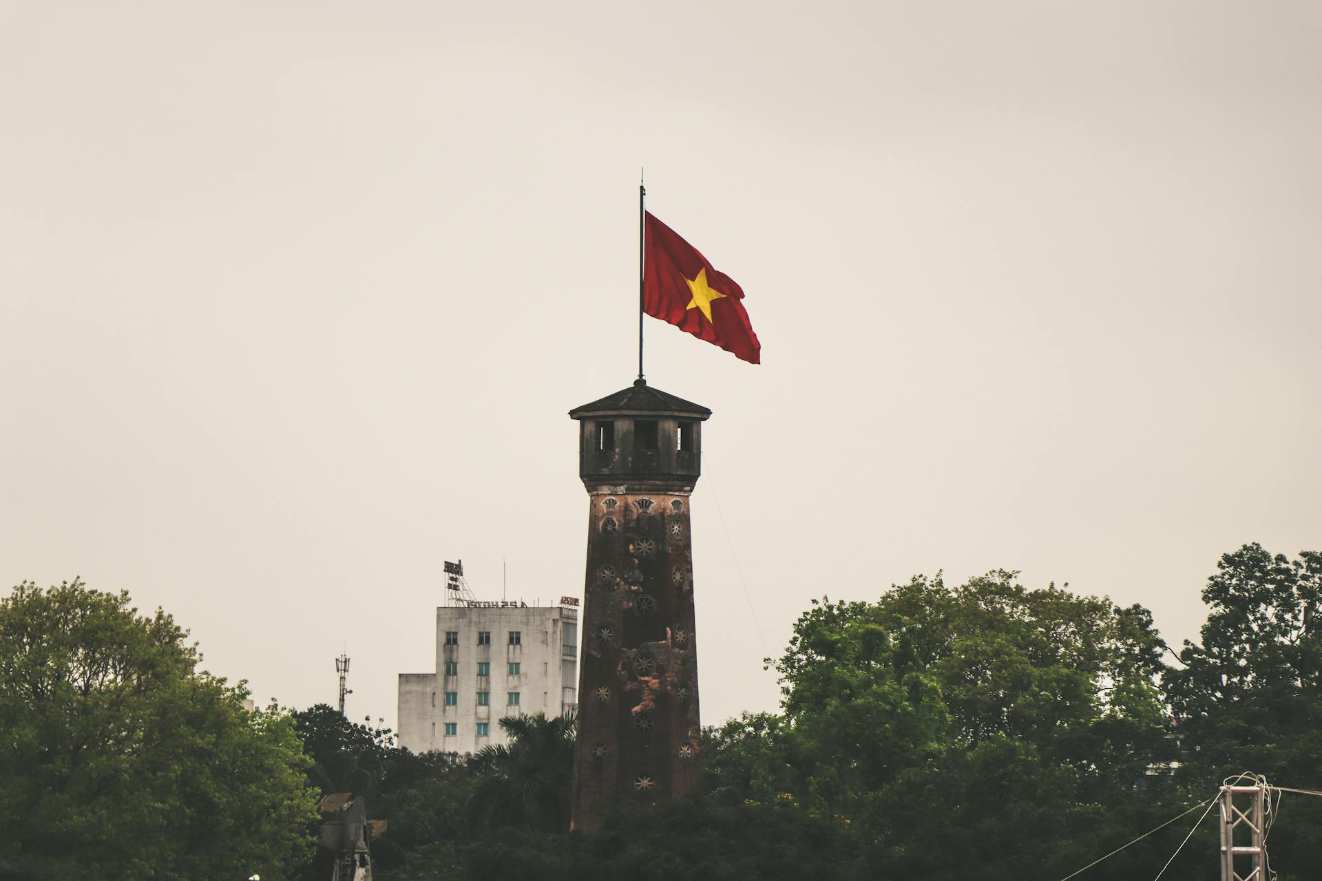 Torre de ladrillo oscuro con la bandera vietnamita roja ondeando en la cima, rodeada de árboles verdes y un edificio blanco al fondo bajo un cielo
