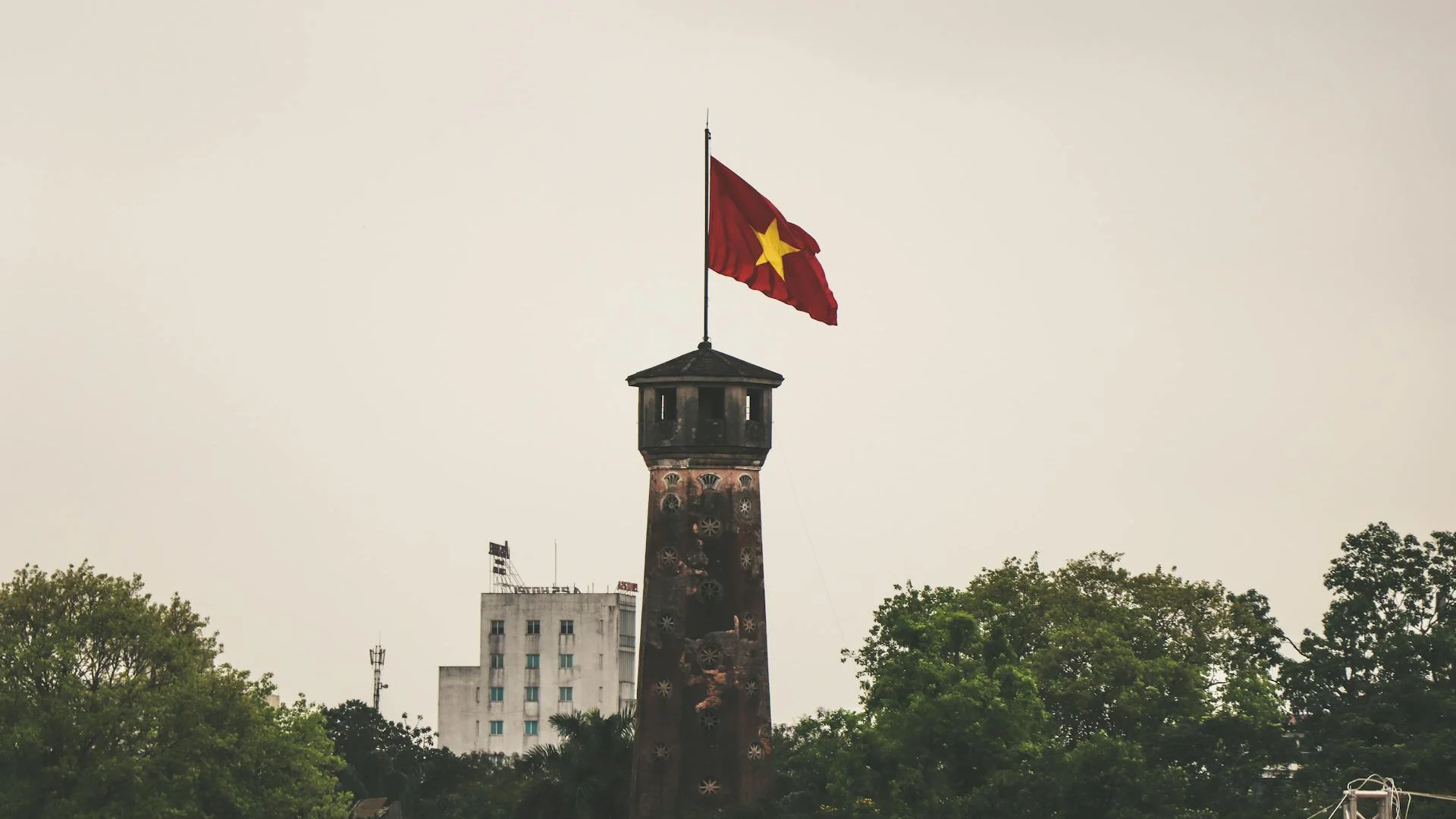 Torre de ladrillo oscuro con la bandera vietnamita roja ondeando en la cima, rodeada de árboles verdes y un edificio blanco al fondo bajo un cielo