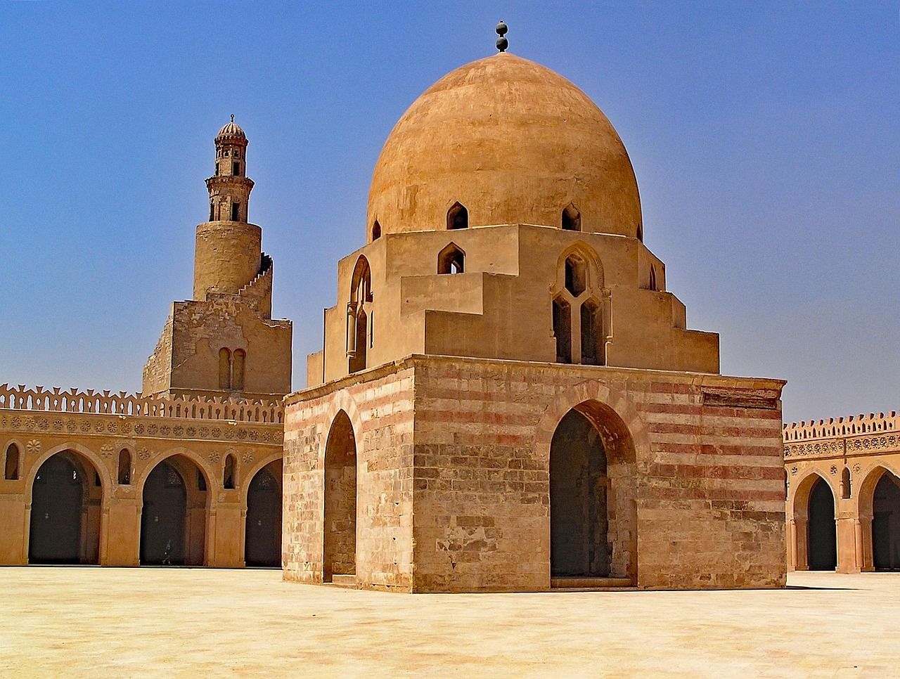 Cúpula y minarete de una mezquita de piedra de tonos tierra bajo un cielo azul claro.