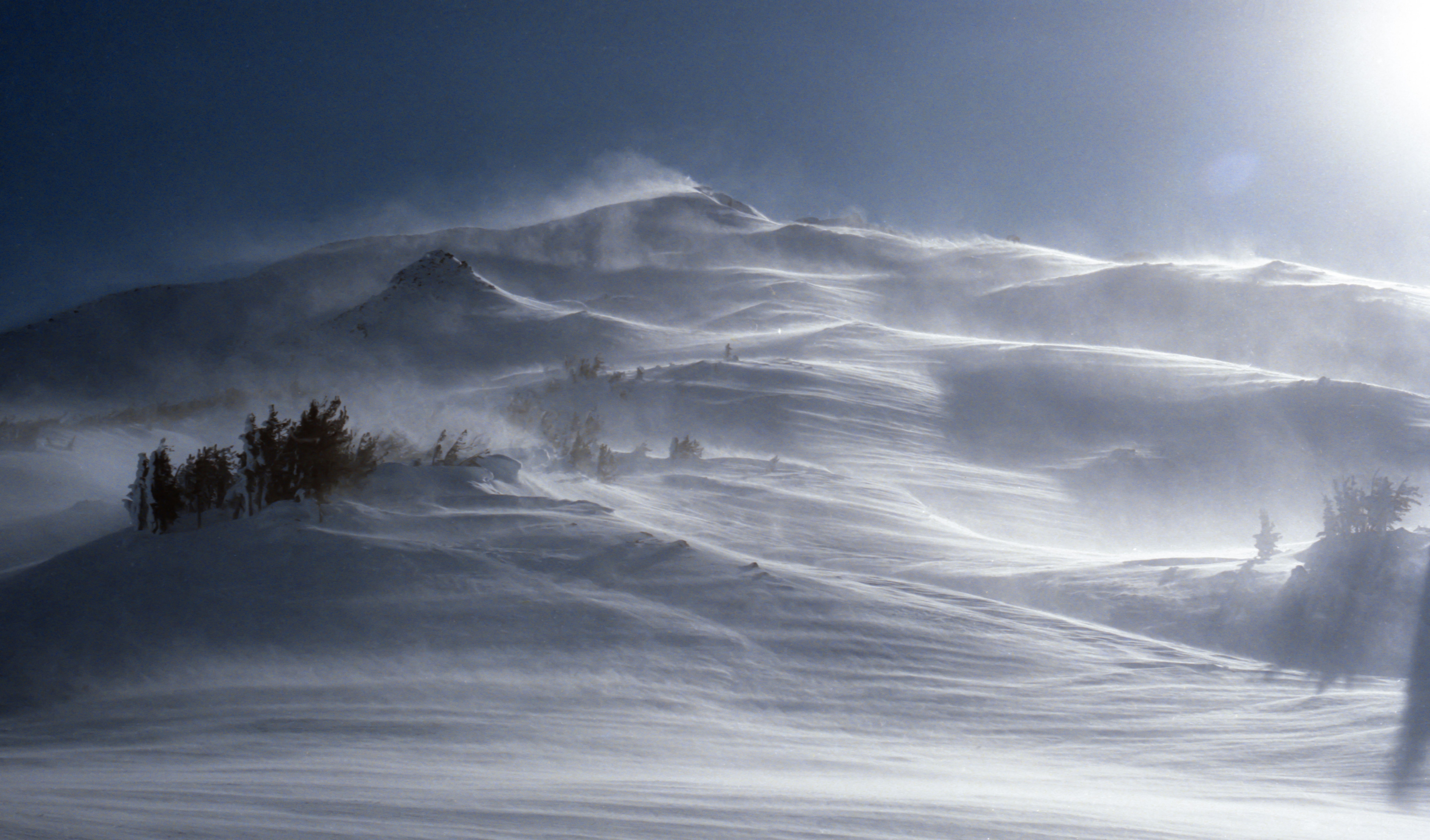 Paisaje montañoso nevado con fuertes ráfagas de viento levantando nieve en el aire.