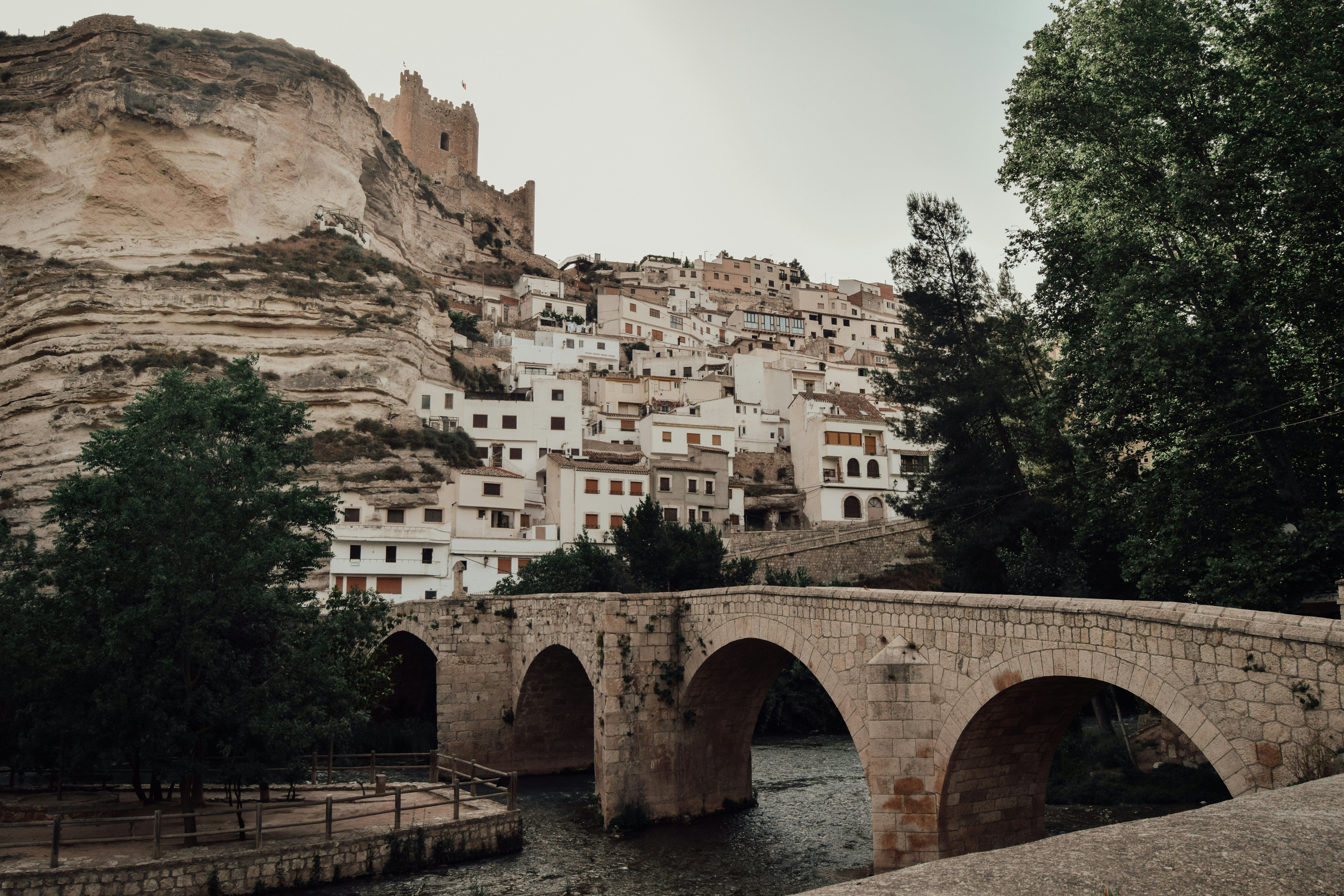 Pueblo blanco con casas en ladera, puente de piedra con arcos sobre río y castillo en la cima.