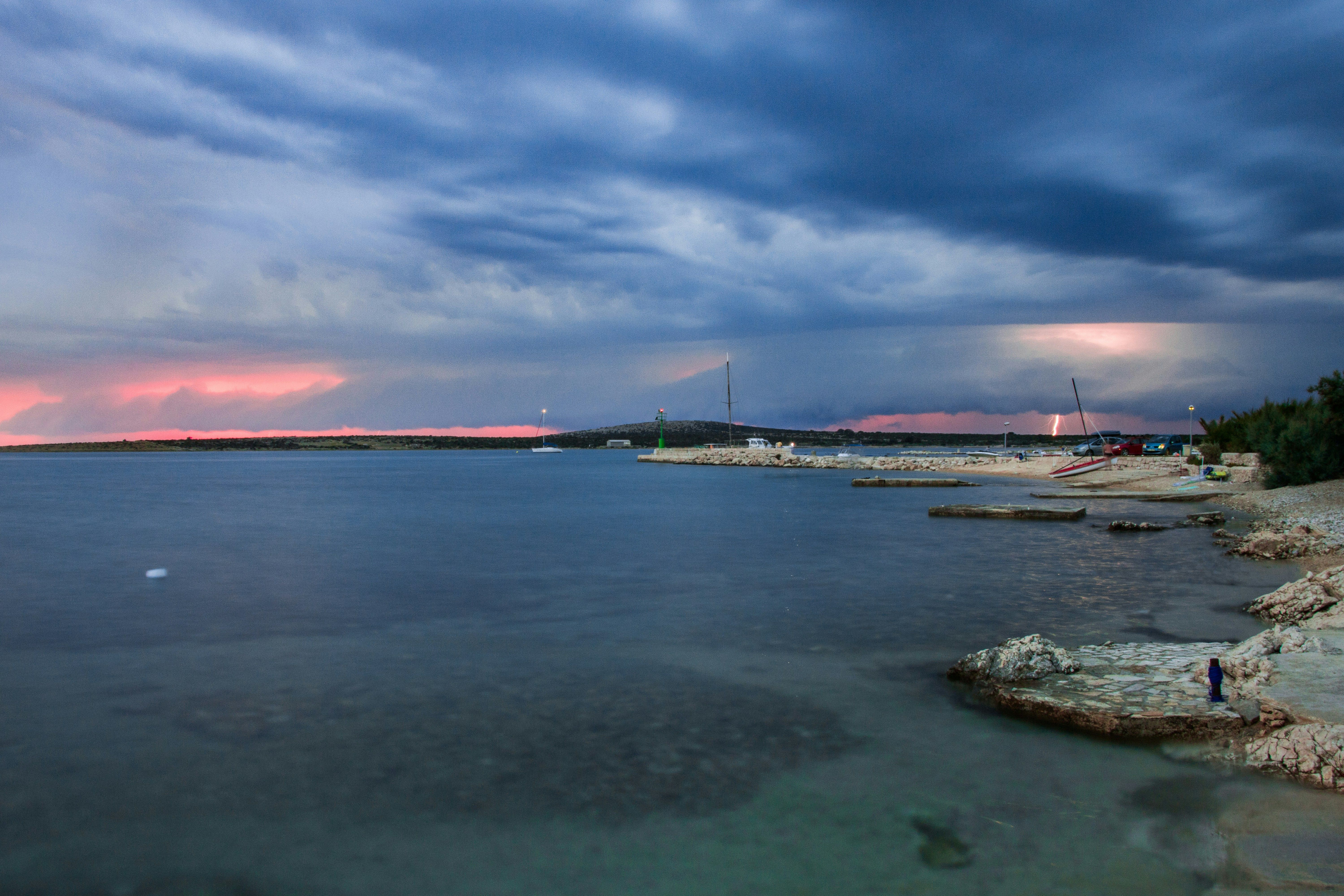 Cielo tormentoso con nubes oscuras y un rayo sobre un mar en calma con una costa rocosa y un puerto.