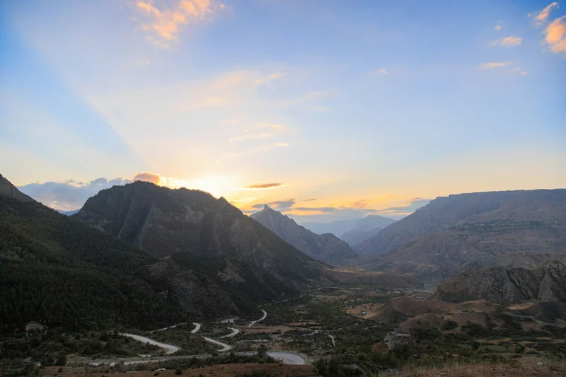 Vista panorámica de un valle montañoso al amanecer, con el sol asomando por detrás de una cumbre.