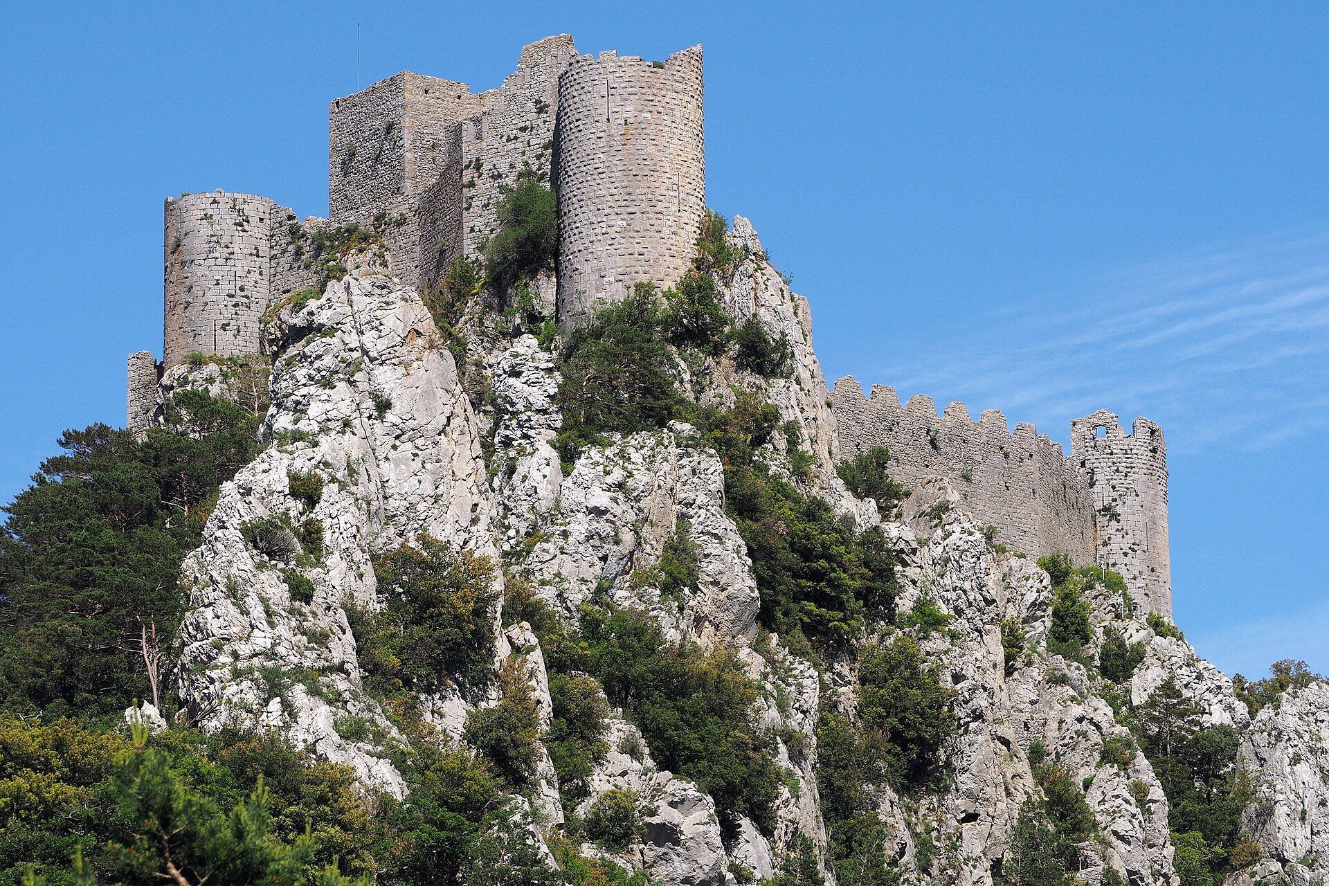 Castillo de piedra medieval en ruinas sobre una cresta rocosa cubierta de vegetación bajo un cielo azul.