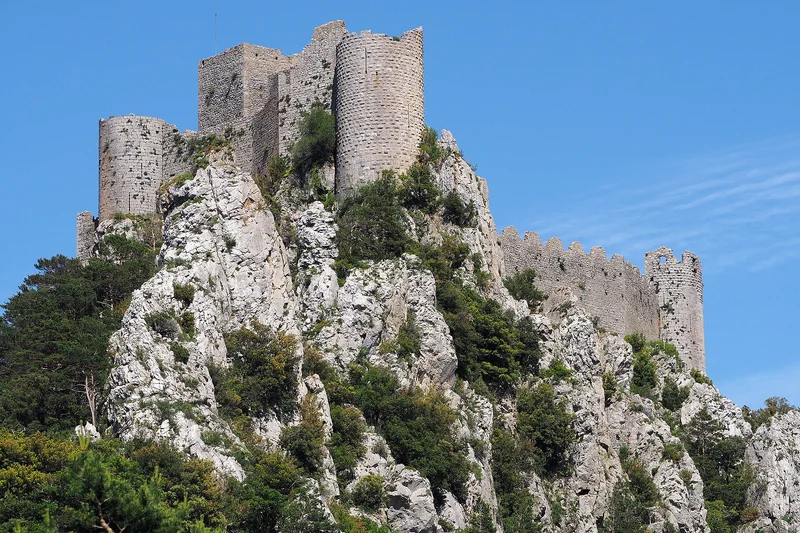 Castillo de piedra medieval en ruinas sobre una cresta rocosa cubierta de vegetación bajo un cielo azul.