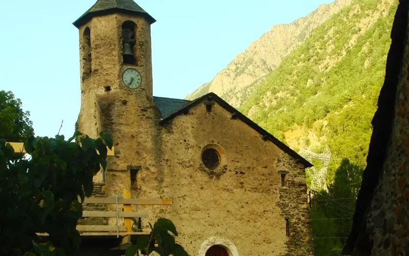 Iglesia antigua de piedra con torre campanario y reloj en Tavascan, rodeada de vegetación y montañas pirenaicas al fondo