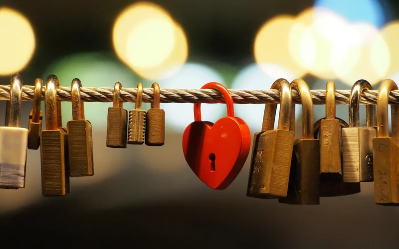 Varios candados de amor colgando de un cable, con uno rojo en forma de corazón destacado, sobre fondo de luces borrosas