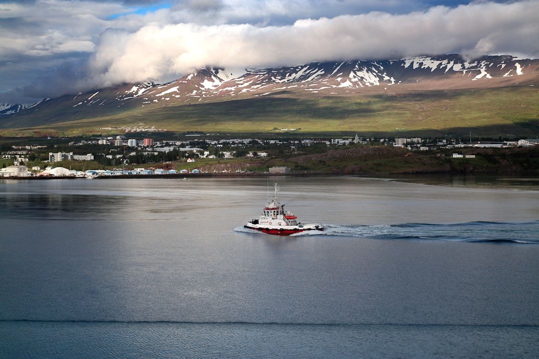 Vista de Akureyri con montañas nevadas y nubes bajo un cielo parcialmente soleado, edificios junto al fiordo y un remolcador rojo navegando en el agua