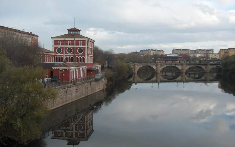 Vista del Puente de Piedra sobre el río Ebro en Logroño, con un edificio rojo a la orilla y reflexiones en el agua bajo un cielo nublado