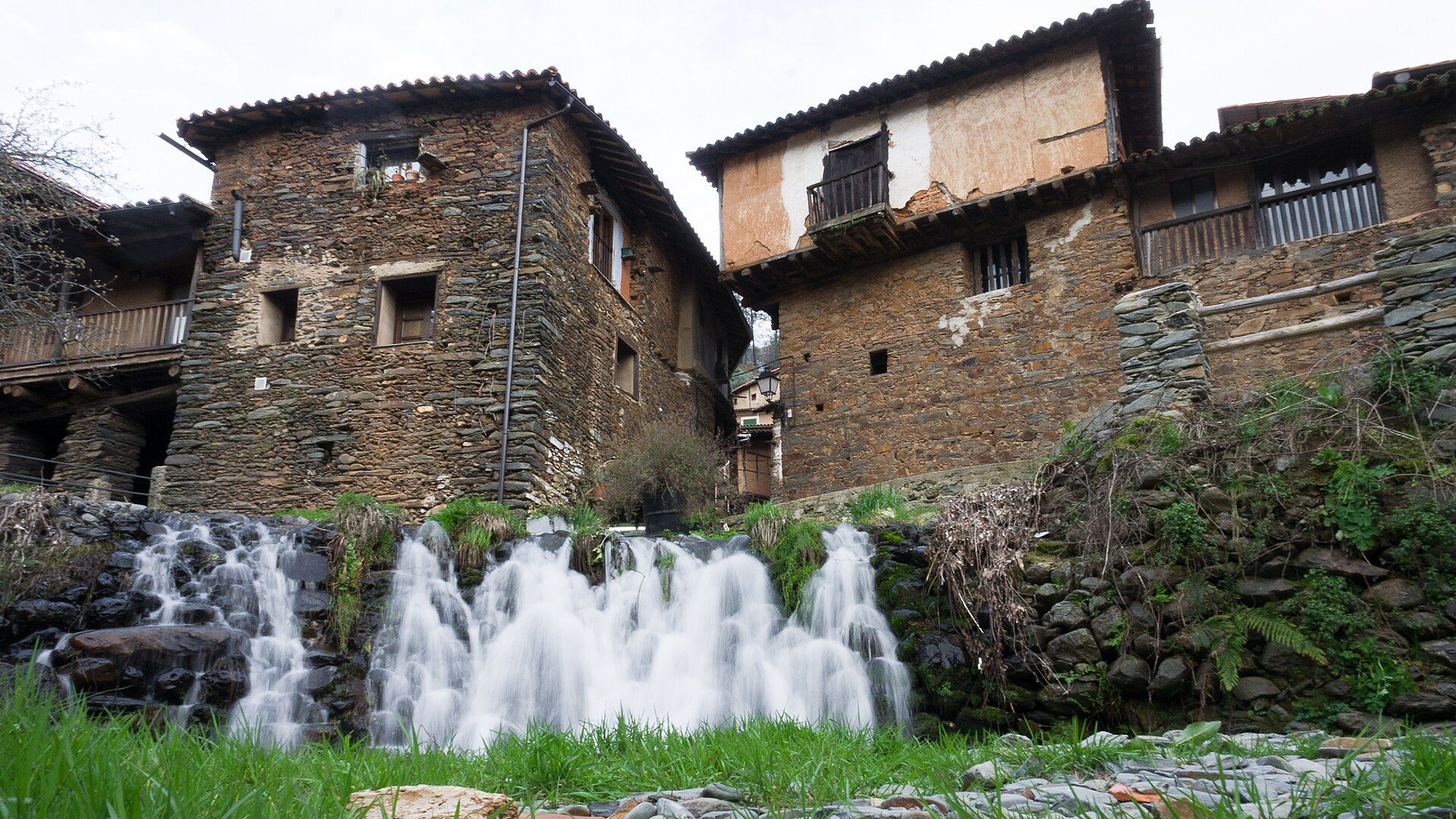Casas de piedra con balcones de madera y tejados de teja, con una cascada en primer plano.