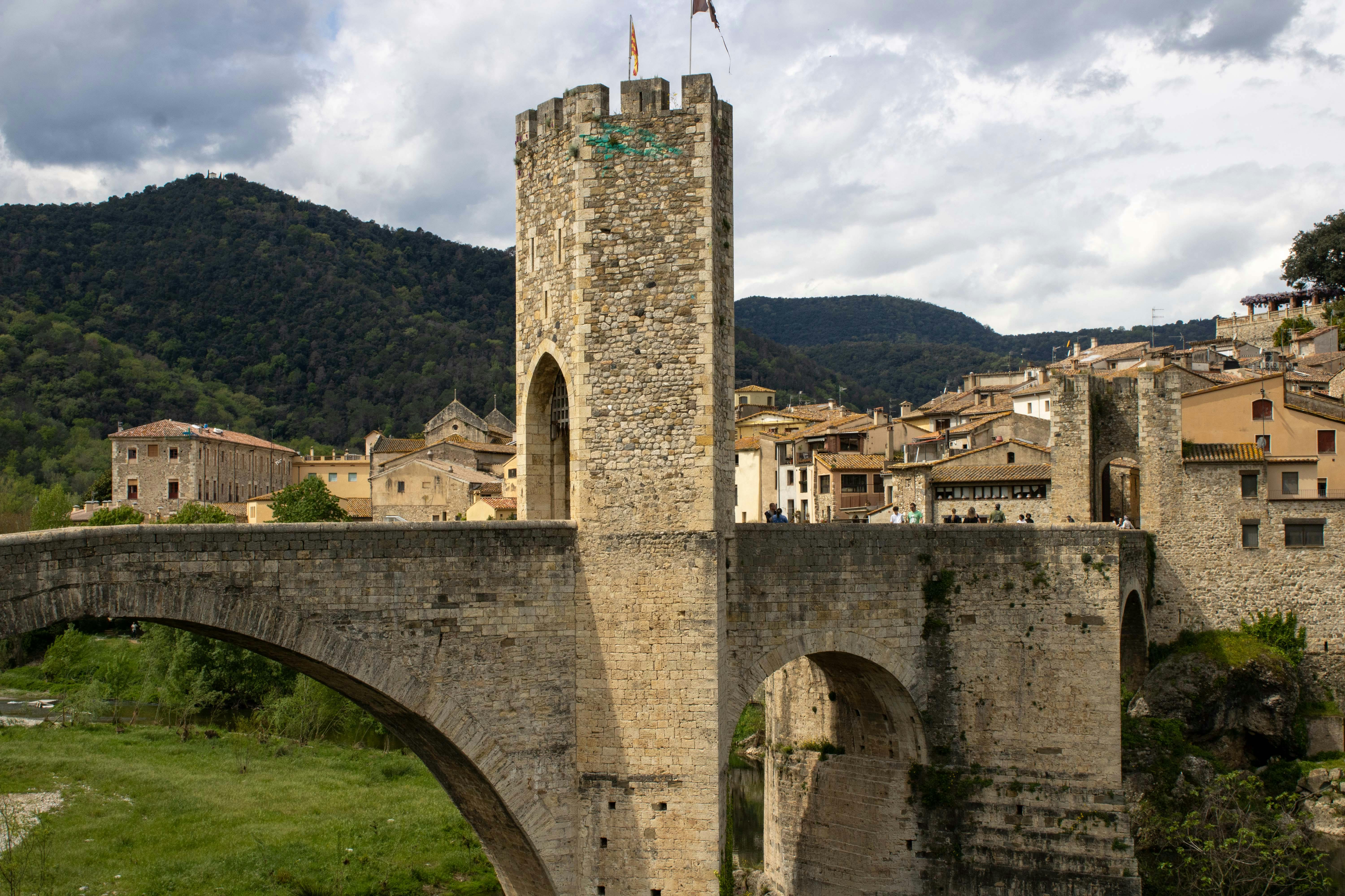 Puente medieval de piedra con torre fortificada y un pueblo al fondo, bajo un cielo nublado.