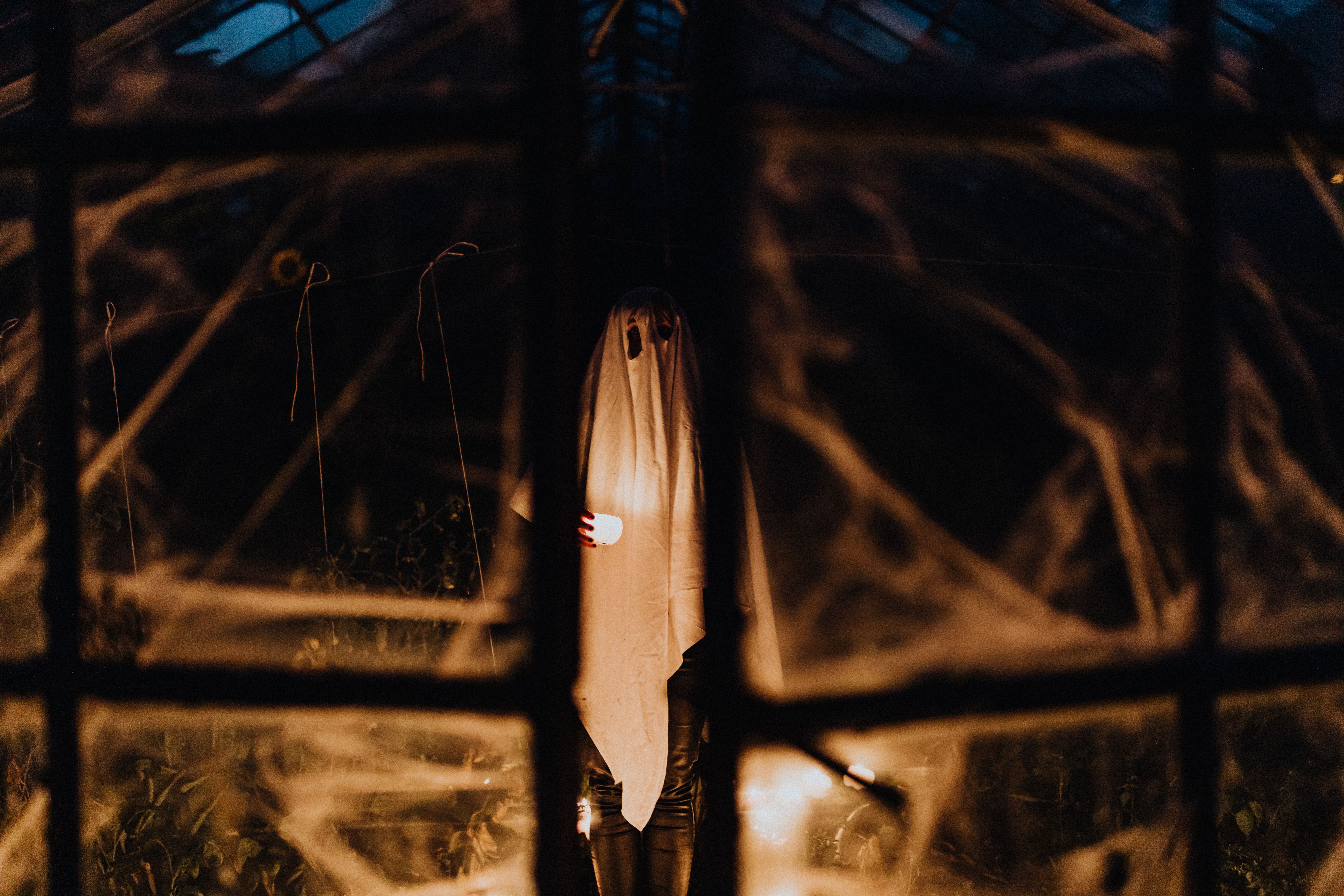 Figura fantasmal cubierta con una sábana blanca y ojos oscuros, sosteniendo una luz, vista a través de una ventana empañada con telarañas.