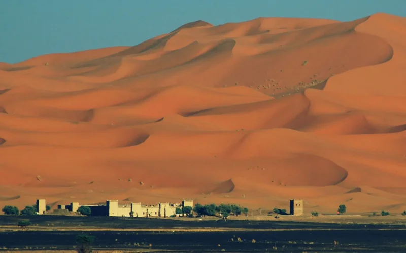 Dunas de arena rojiza se curvan en el desierto de Merzouga, con edificios tradicionales blancos y palmeras en primer plano bajo un cielo azul