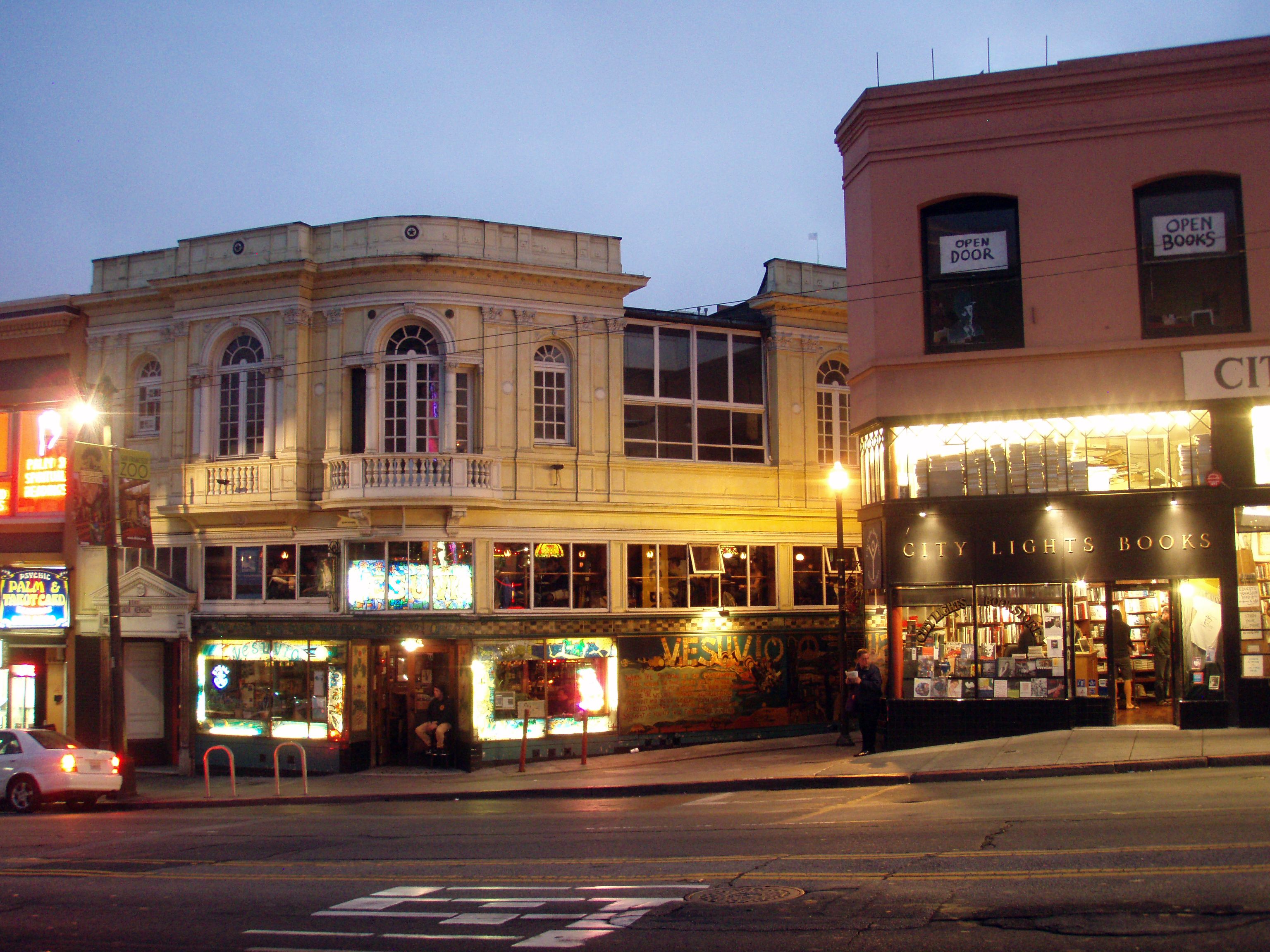 Fachada iluminada de la librería City Lights Books al anochecer, junto a un edificio de color crema con el bar Vesuvio en la planta baja.