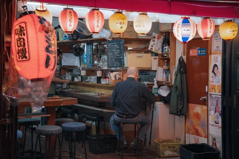 Hombre sentado de espaldas en una barra de bar japonés con farolillos y carteles publicitarios.