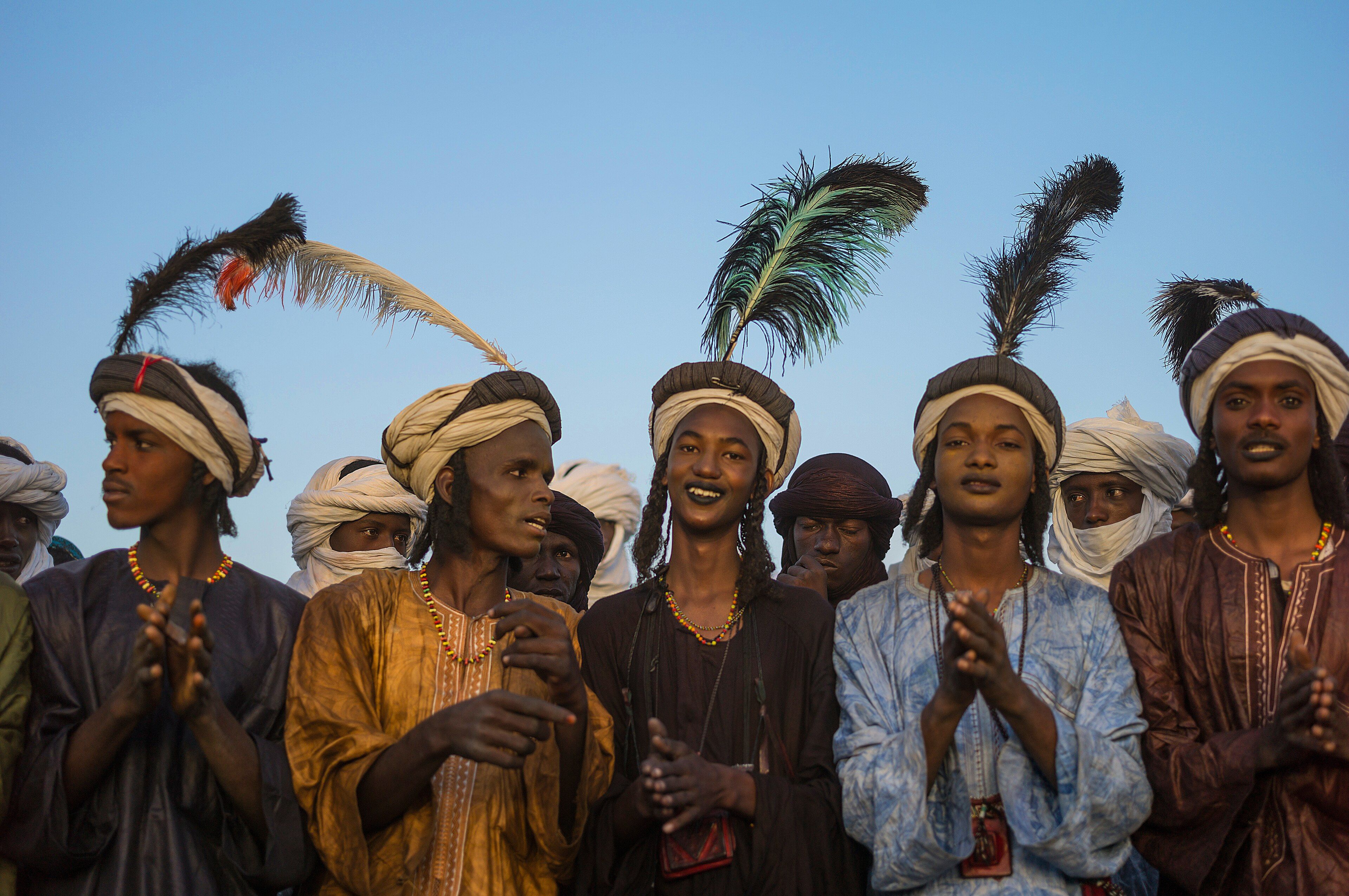 Fila de jóvenes con turbantes, plumas negras y túnicas de colores que dan palmas bajo un cielo azul, con hombres con turbante blanco detrás.