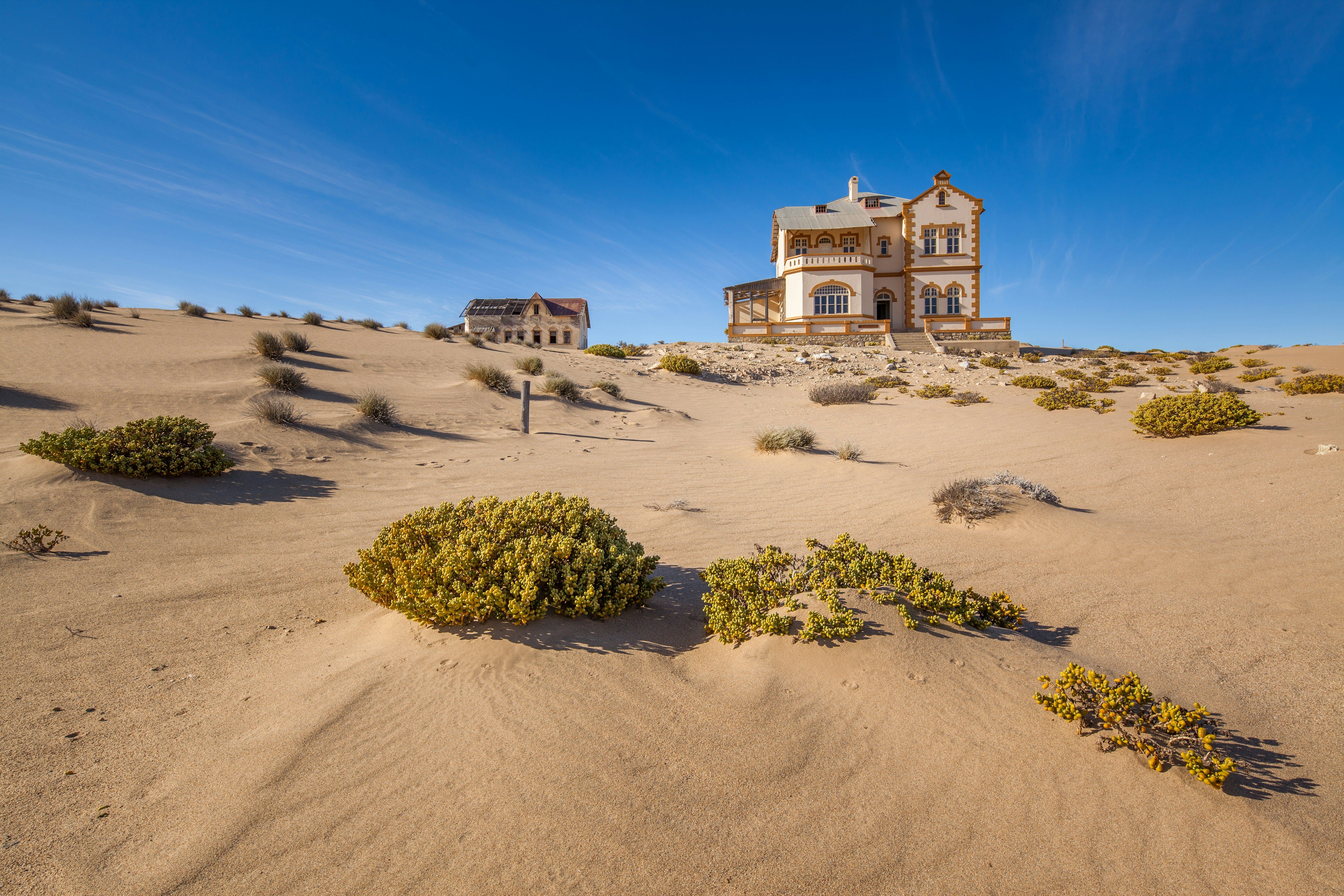 Edificios abandonados de estilo colonial rodeados por dunas de arena y vegetación desértica bajo un cielo azul.
