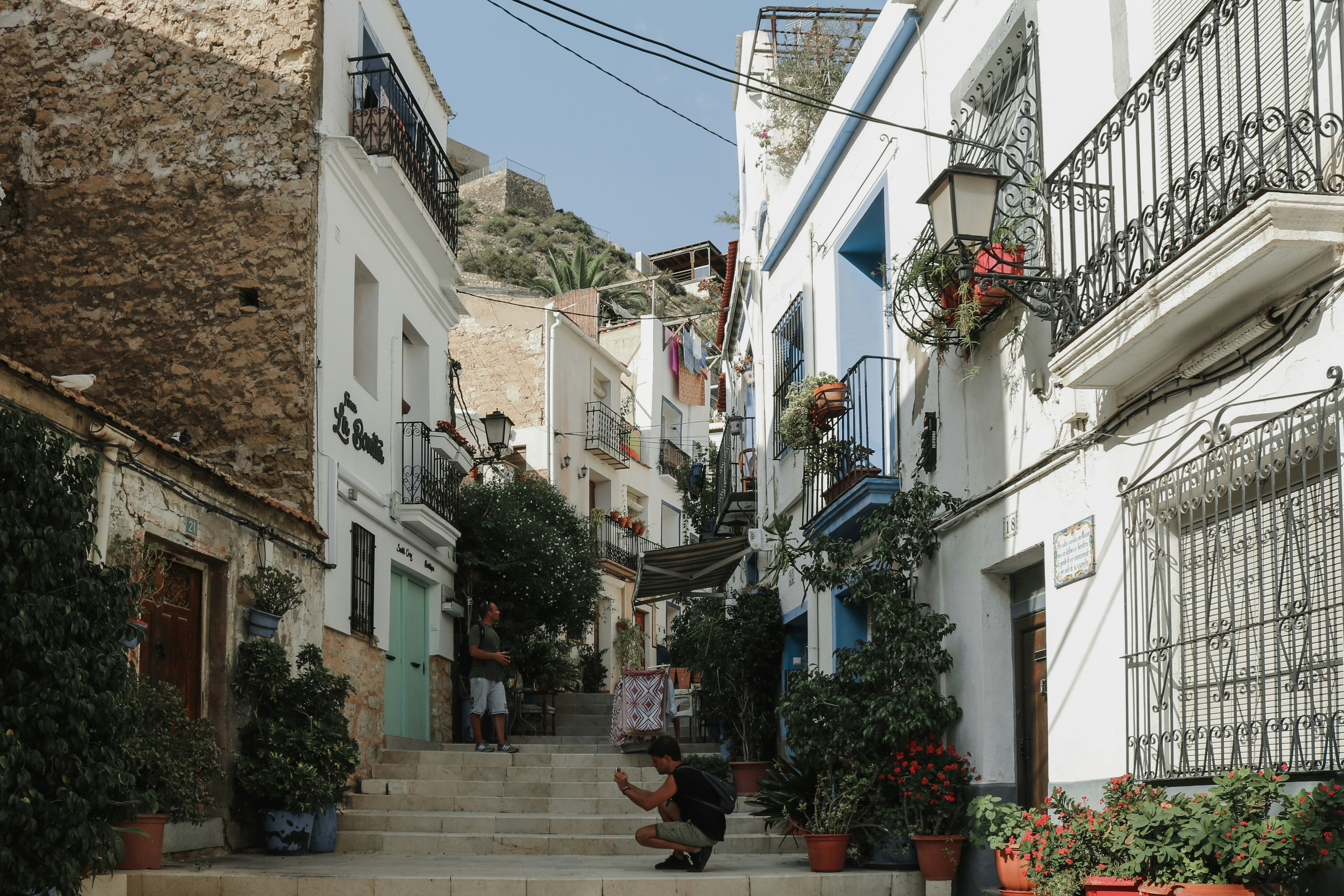Calle empedrada estrecha con casas blancas y balcones, plantas en macetas y un hombre agachado.