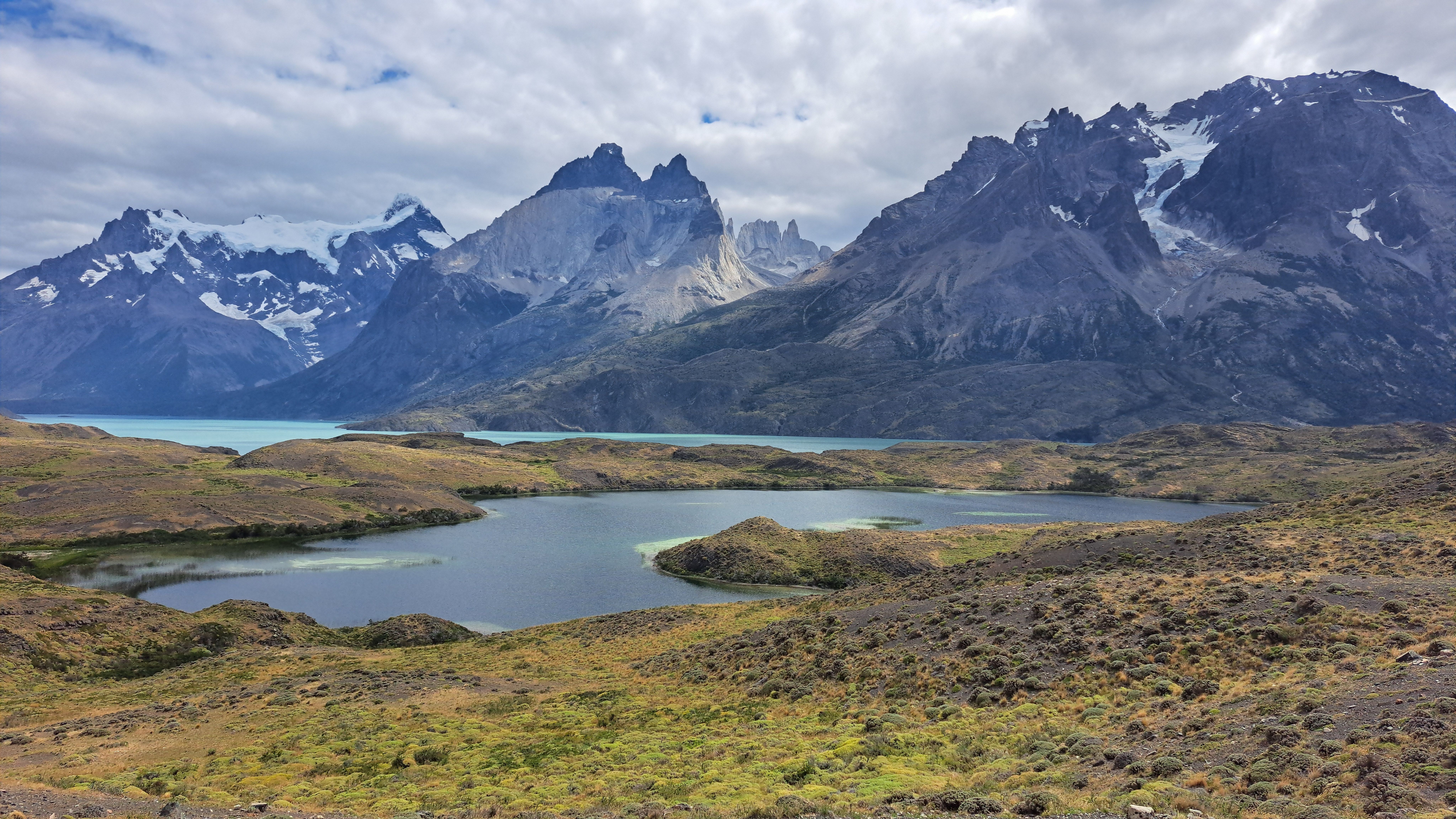 Paisaje de montaña con picos nevados, lagos de agua azul y vegetación de estepa bajo un cielo nublado.